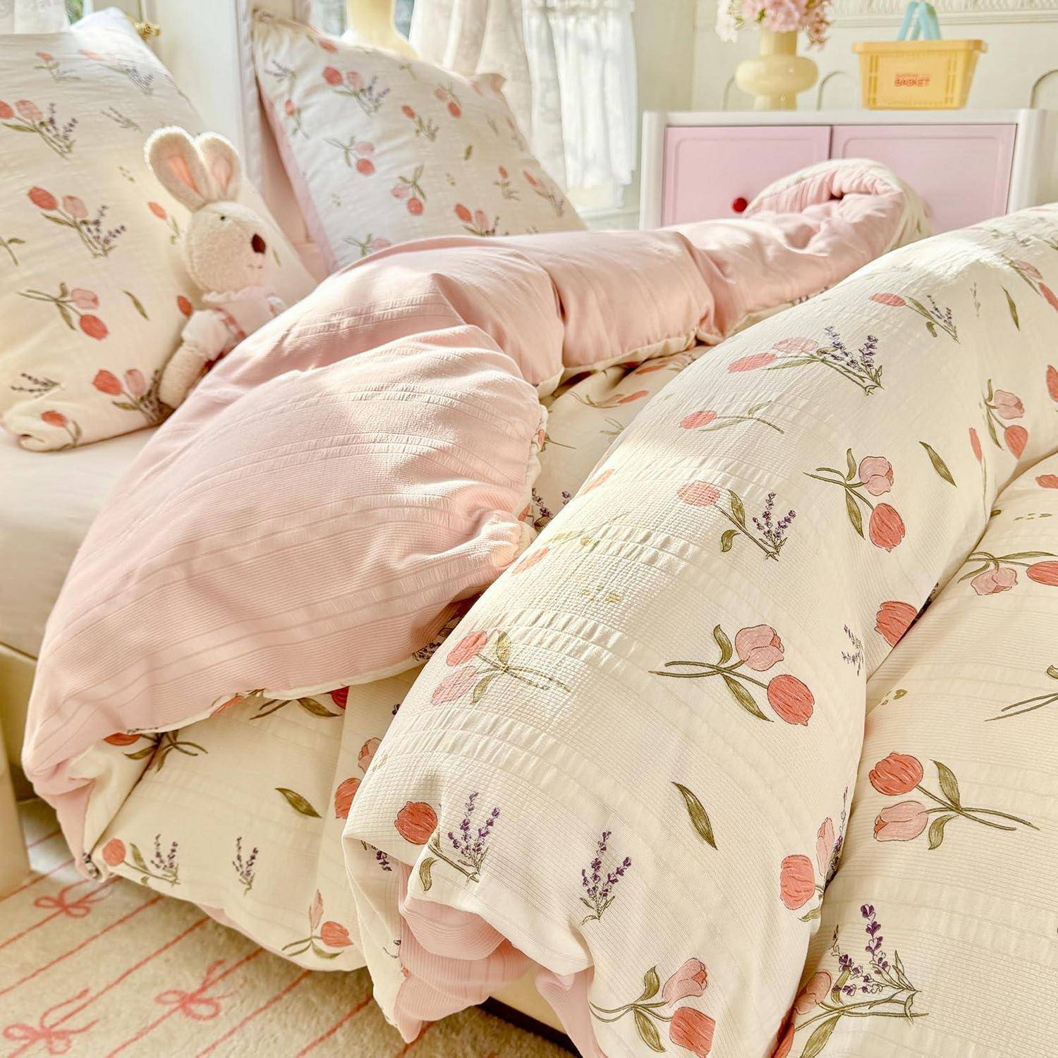 Blumenbettwäsche mit einer rosa Decke und Hasenplüsch auf einem ordentlich gemachten Pastellbett in einem hellen Schlafzimmer.