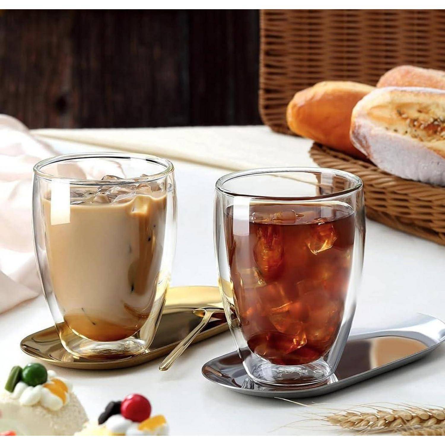 Zwei Glastassen mit Eiskaffee und Milchtee auf Untertassen neben Brötchen auf einem Frühstückstisch.