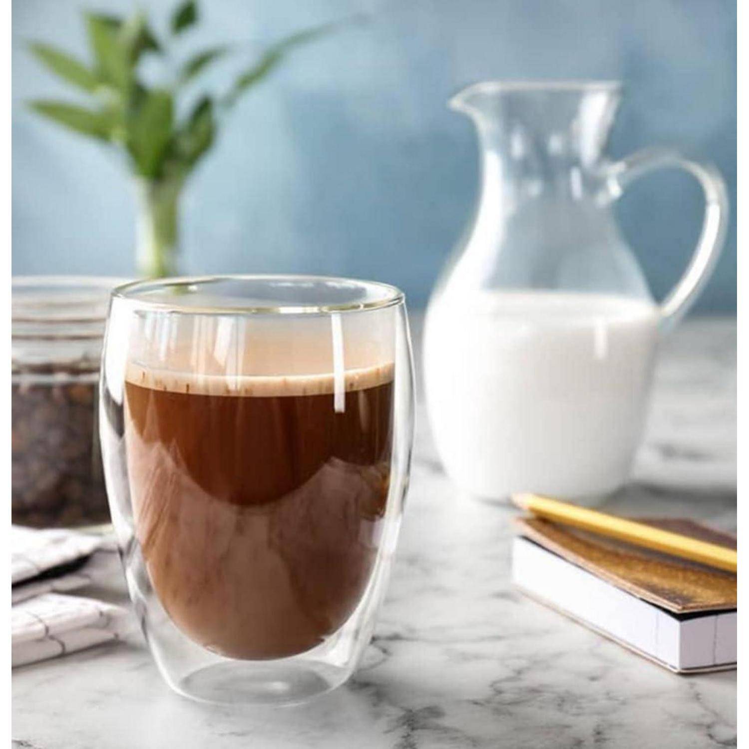 Doppelwandiges Glaskaffee auf einem Marmortisch, mit einer Milchkanne, einem Notizbuch und einer Pflanze im Hintergrund.