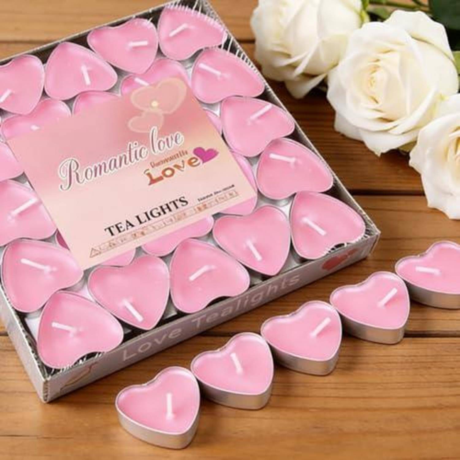 Schachtel mit rosa herzförmigen Teelichtern mit der Aufschrift "Romantische Liebe"" auf einer Holzoberfläche neben weißen Rosen.