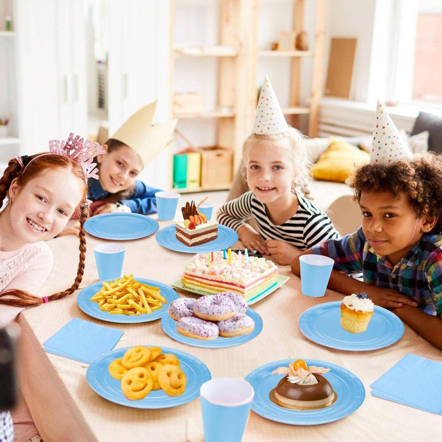 Vier Kinder in Partyhüten lächeln um einen Geburtstagstisch mit Kuchen, Keksen, Pommes und blauen Bechern.