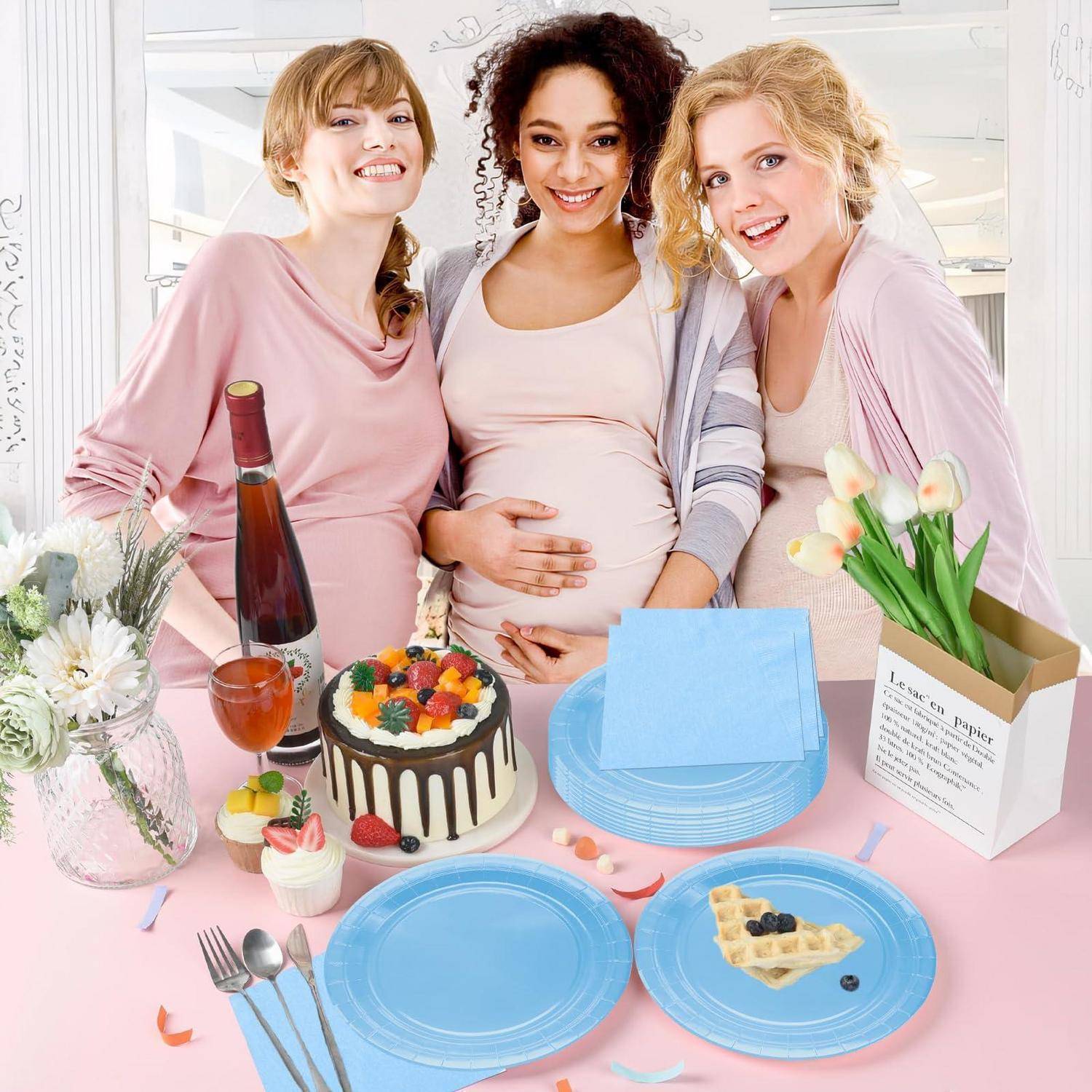 Drei lächelnde schwangere Frauen posieren an einem Babyduschtisch mit Kuchen, blauen Tellern, Tulpen, Getränken und Geschenktüten.