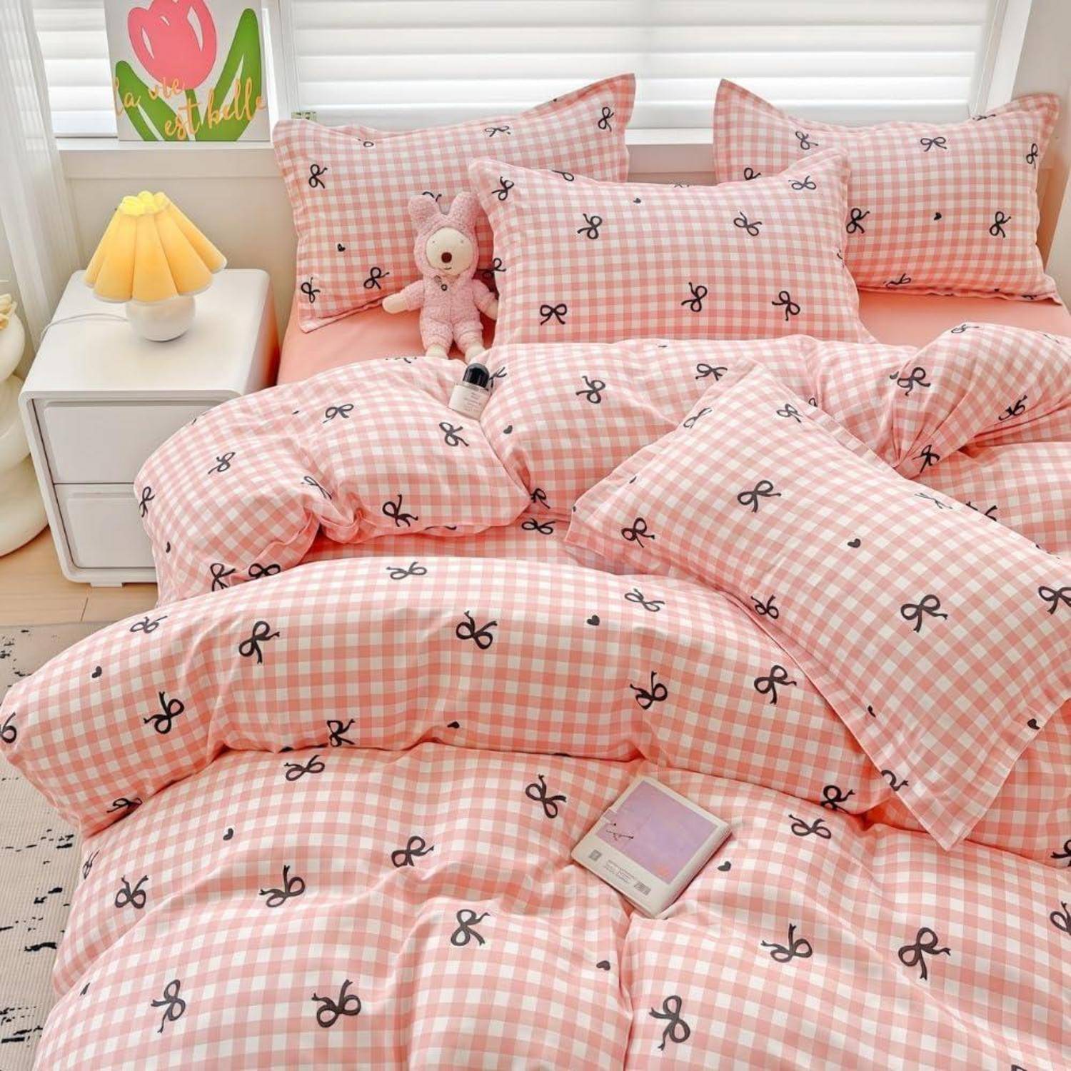 Rosa Gingham-Bettwäsche mit schwarzem Schleifenmuster auf einem ordentlich gemachten Bett, mit einem kleinen rosa Teddybär, einer Nachttischlampe und einem Buch auf der Tagesdecke.