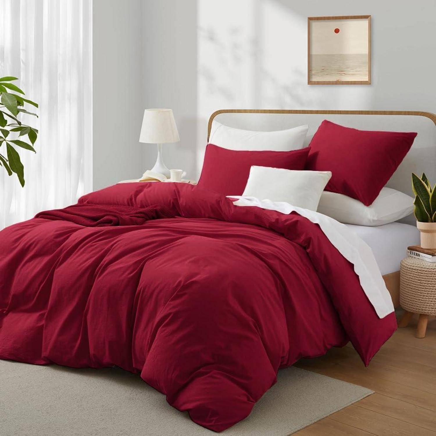 Ordentlich gemachtes Bett mit tiefroter Bettwäsche und weißen Kissen in einem hellen, minimalistischen Schlafzimmer mit einer Nachttischlampe und Pflanzen.