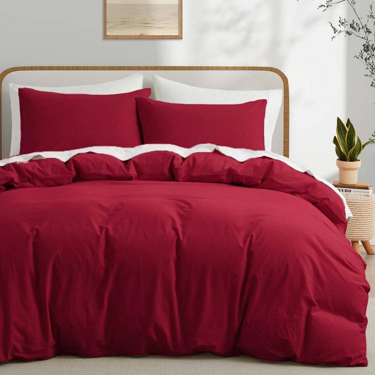 Ein Bett mit tief roter Bettwäsche und Kissen in einem minimalistischen Schlafzimmer, mit einer Pflanze und gerahmter Kunst im Hintergrund.