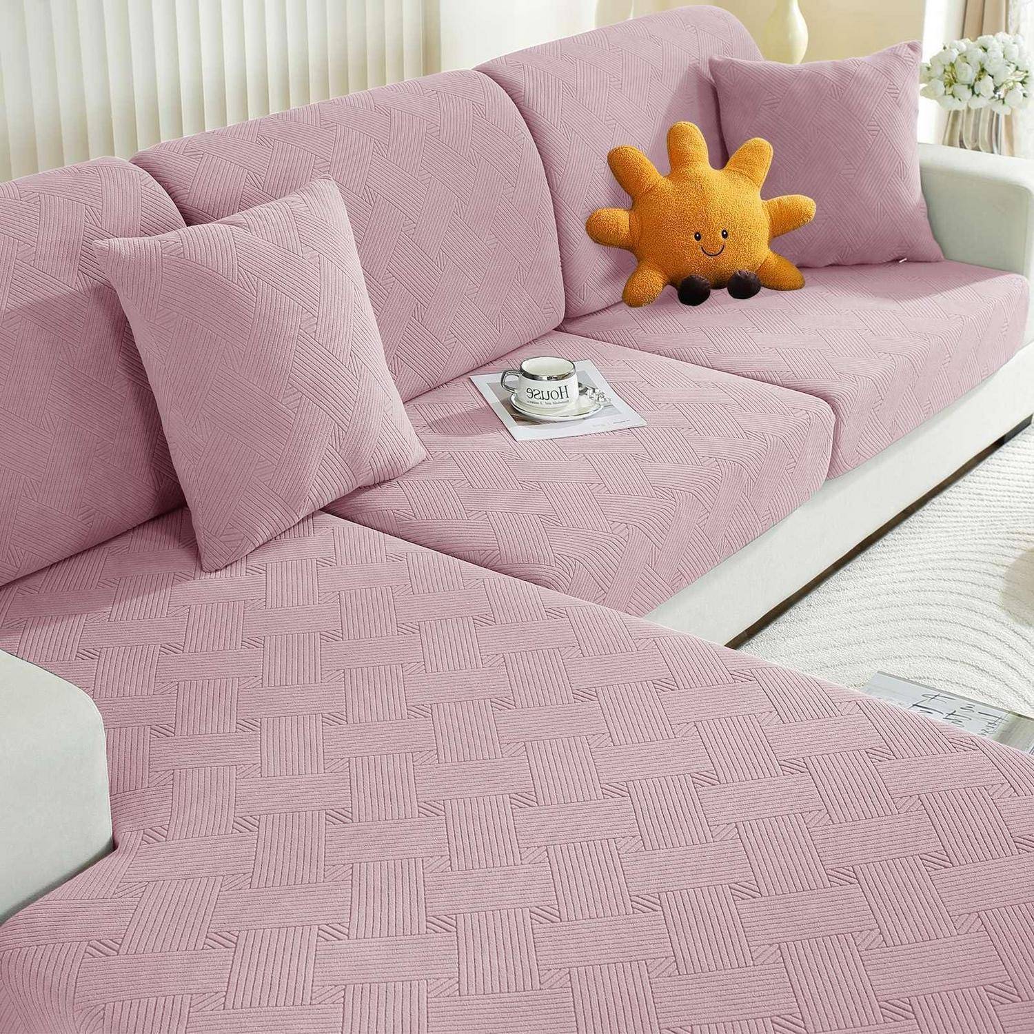Rosa L-förmiges Sofa mit strukturiertem Sofabezug, zwei passende Kissen, ein lachendes orangefarbenes Sonnenplüschtier und eine Kaffeetasse auf einem Tablett.