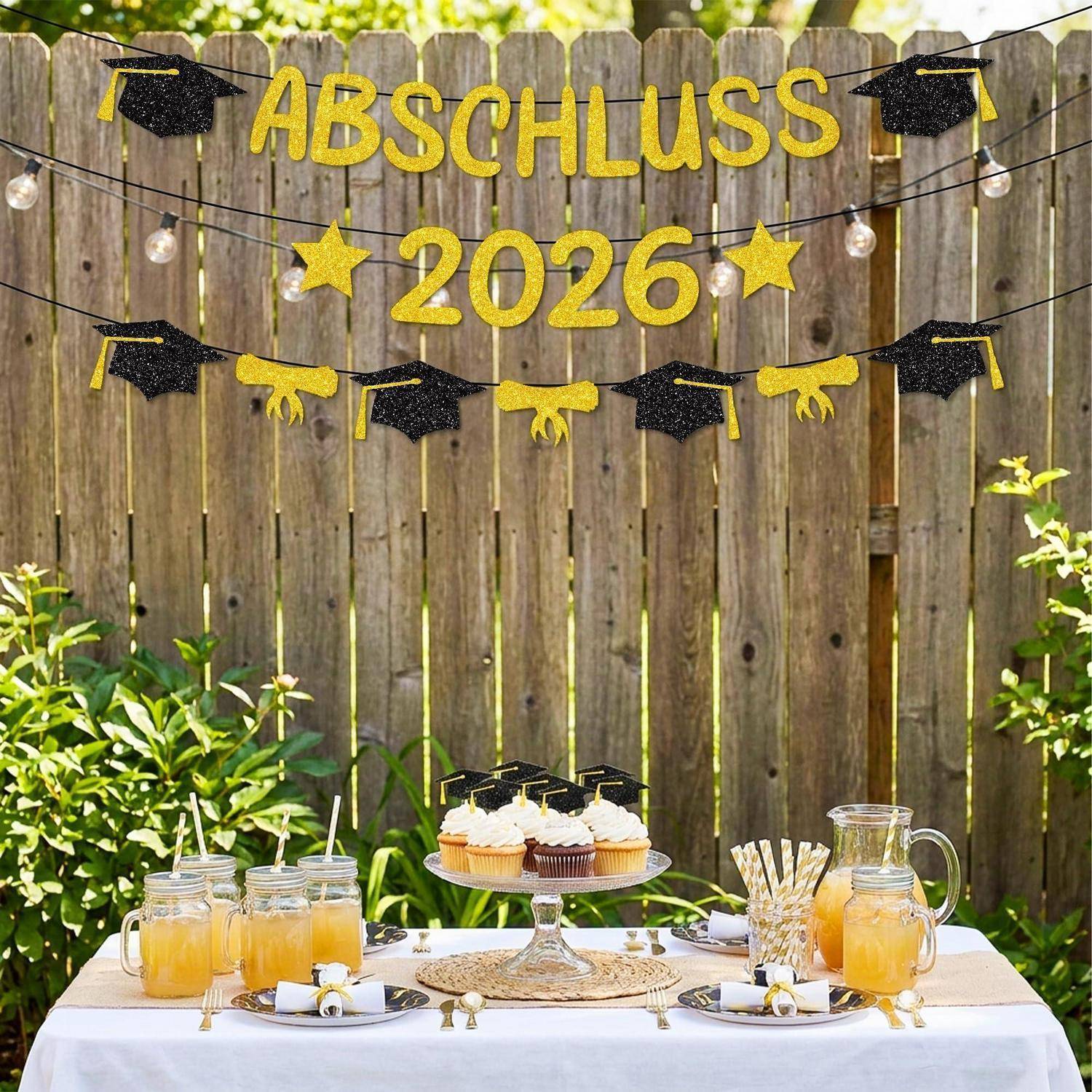Outdoor-Abschlussfeier-Setup mit „ABSCHLUSS 2026"-Banner, Absolventenhut-Girlanden und einem Dessert- und Getränketisch vor einem Holzzaun.