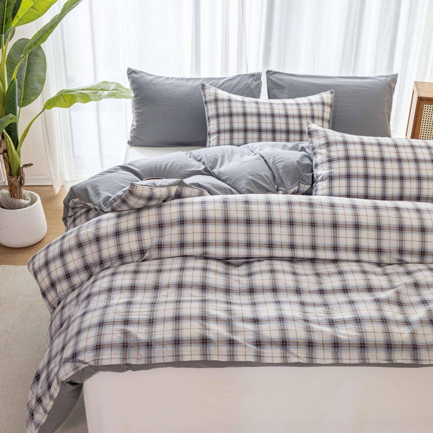 Ordentlich gemachtes Bett mit grauer und karierter Bettwäsche und Kissen in einem hellen Schlafzimmer neben einer Topfpflanze und Fenstervorhängen.