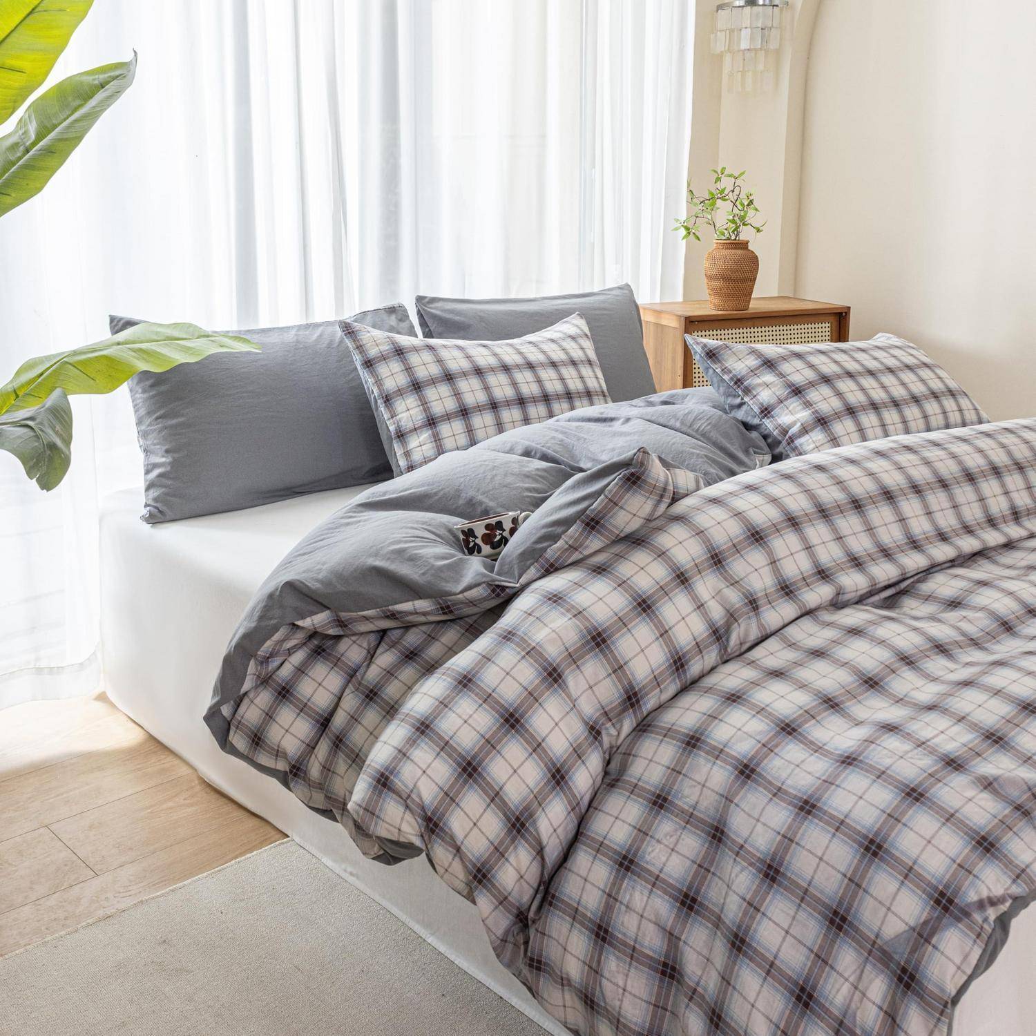 Ein ordentlich gemachtes Bett mit grauer und karierter Bettwäsche in einem hellen Schlafzimmer neben einem kleinen hölzernen Nachttisch und einer Topfpflanze.