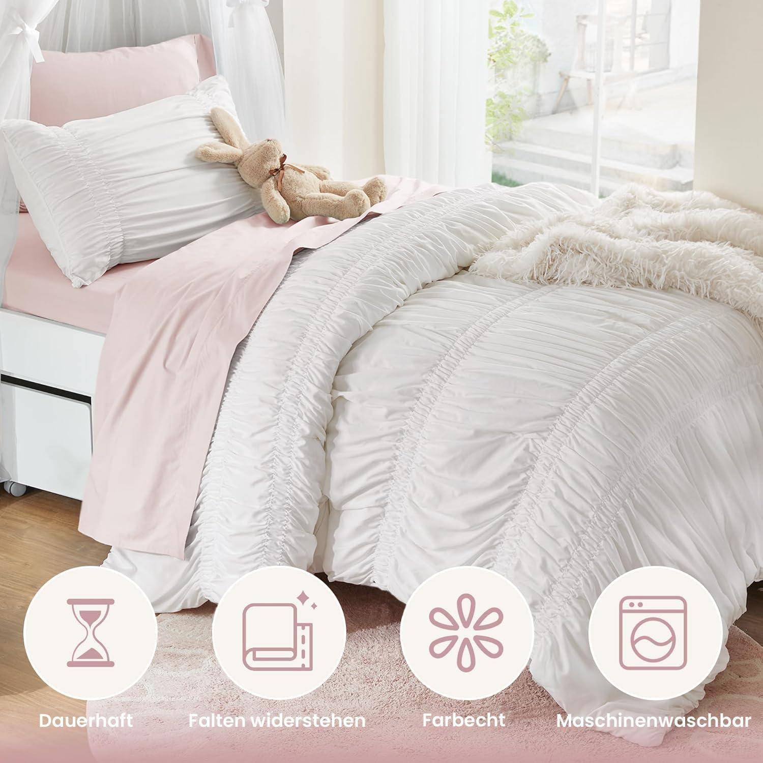 Rosa-weiße gerüschte Bettdecke auf einem ordentlich gemachten Bett mit einem Teddybär, darunter beschriftete Symbole auf Deutsch: 'Dauerhaft', 'Falten widerstehen', 'Farbecht', 'Maschinenwaschbar'.