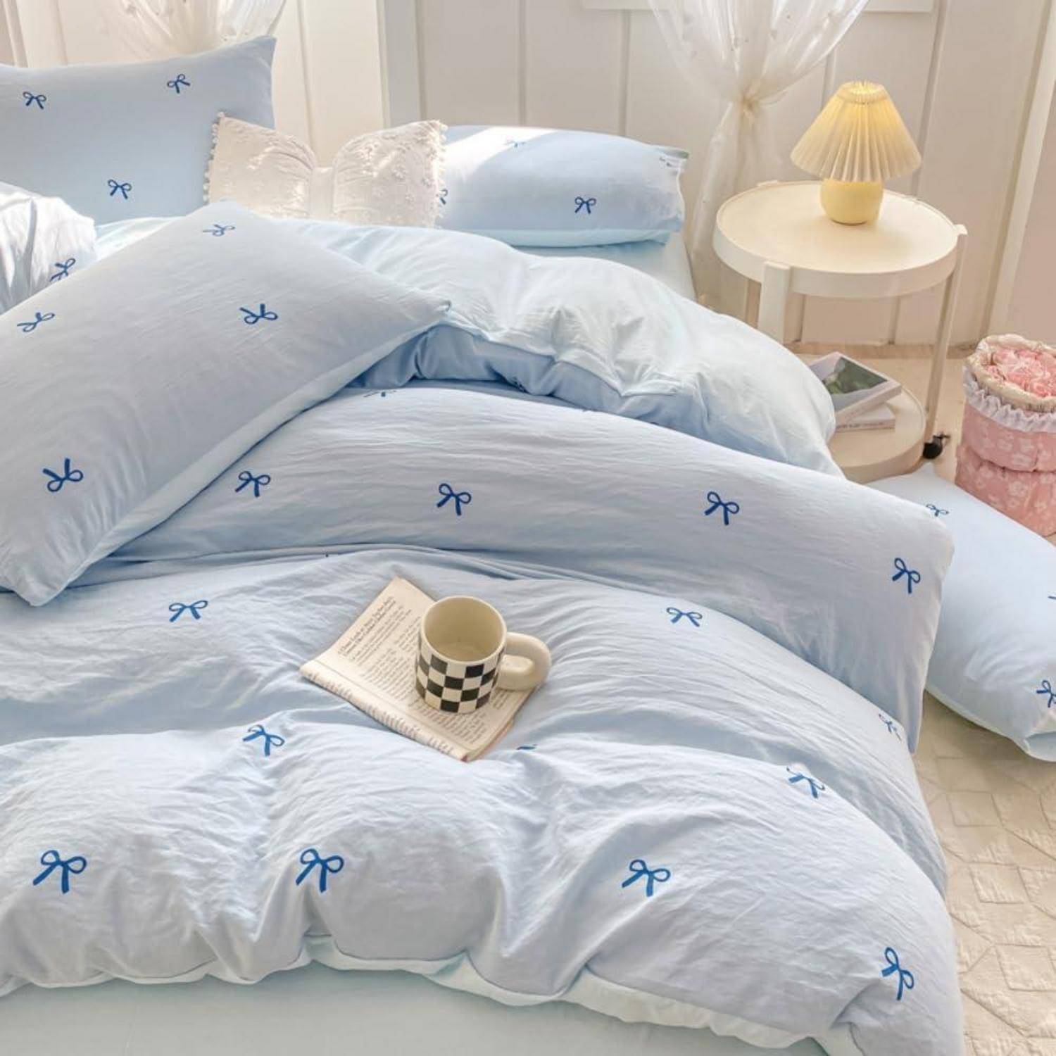 Hellblaues Bett mit Bettwäsche im Schleifenmuster, eine karierte Tasse auf einem Buch und ein Nachttisch mit einer Lampe in einem hellen Schlafzimmer.