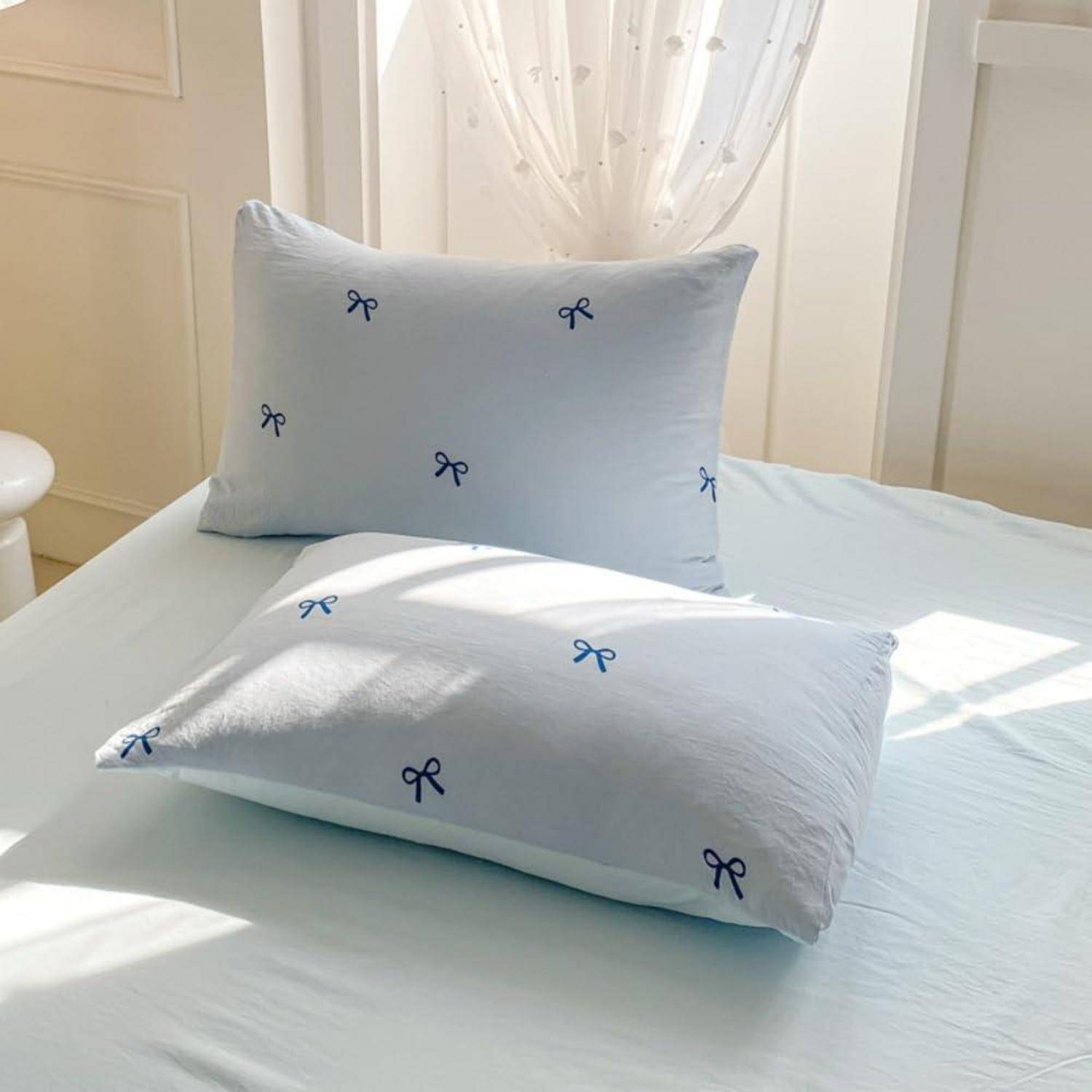 Zwei hellblaue Kissen mit kleinen dunkelblau gemusterten Schleifen auf einem ordentlich gemachten Bett in einem sonnigen Schlafzimmer.
