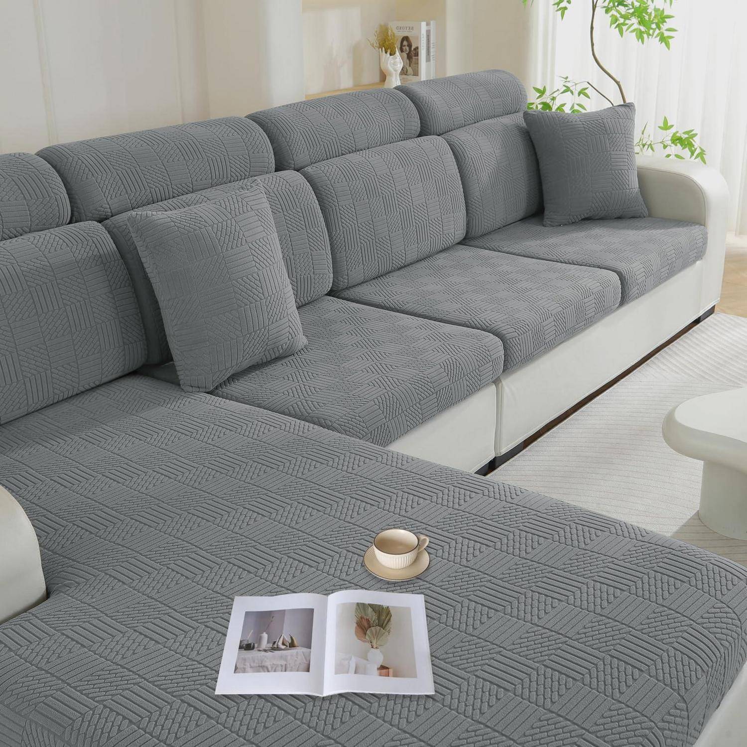 Graues L-förmiges Ecksofa mit strukturierten Schonbezügen und Wurfkissen in einem hellen Wohnzimmer, mit einer offenen Zeitschrift auf der Chaiselongue.
