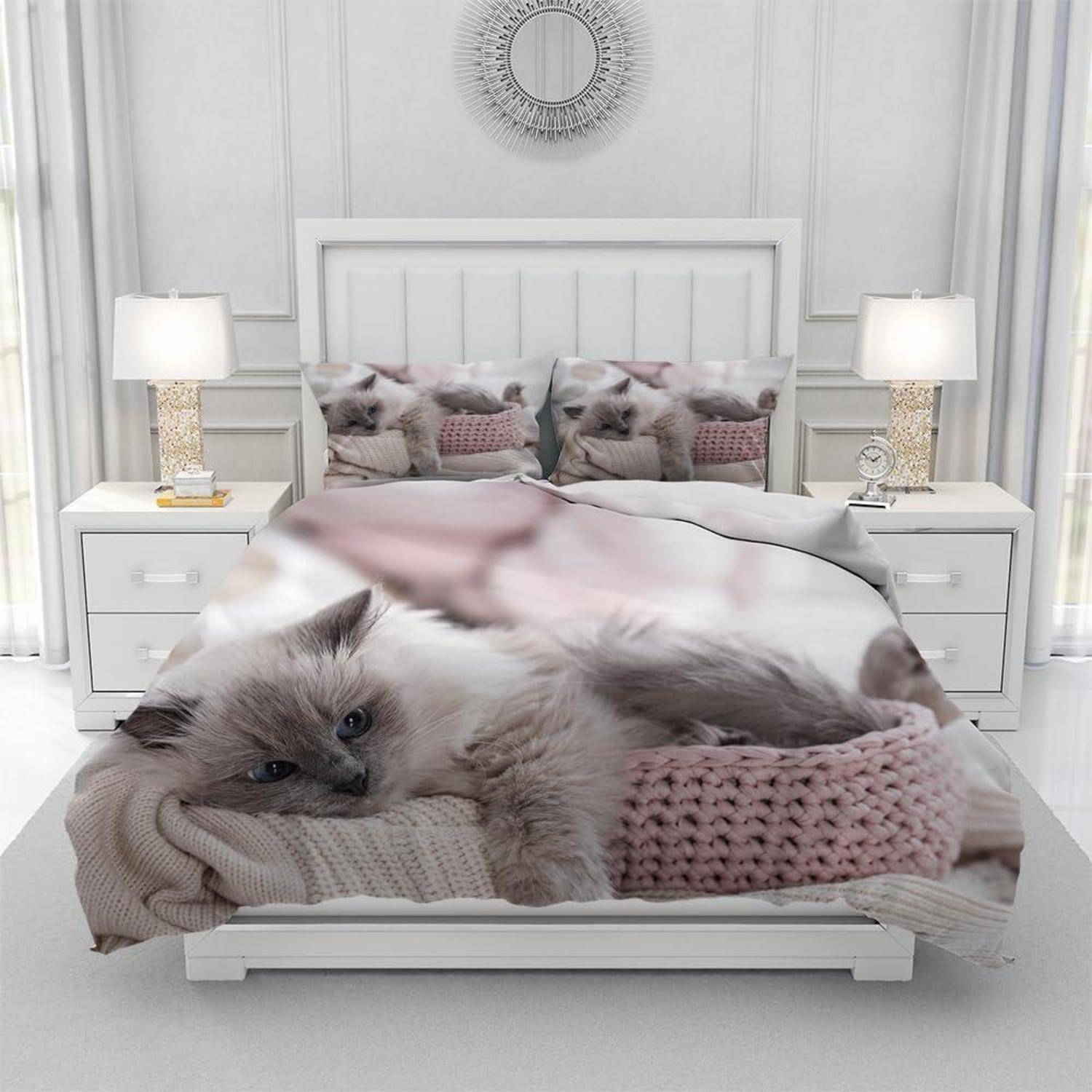 Fluffige graue Katze, die auf einem Bett in einem hellen, weißen Schlafzimmer mit passenden Nachttischlampen und gespiegelten Kissen liegt.