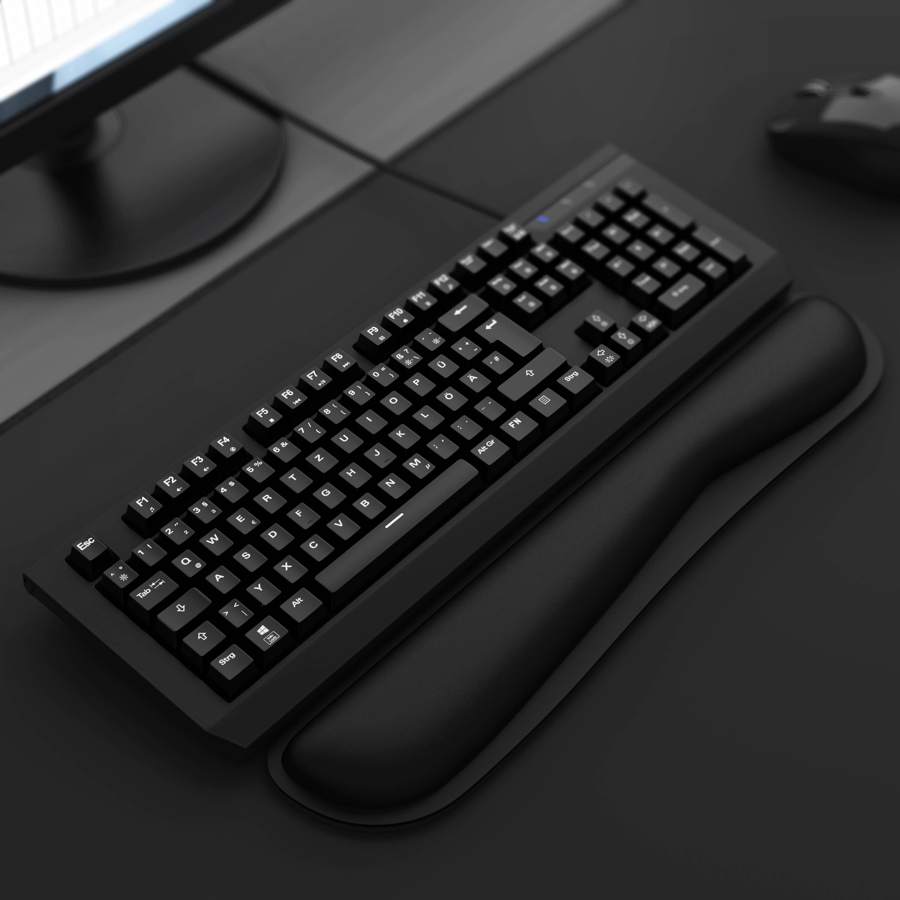 Schwarze mechanische Tastatur mit erhabenen Tasten und Handgelenkstütze auf einem dunklen Schreibtisch, mit teilweise sichtbarem Monitor und Maus.