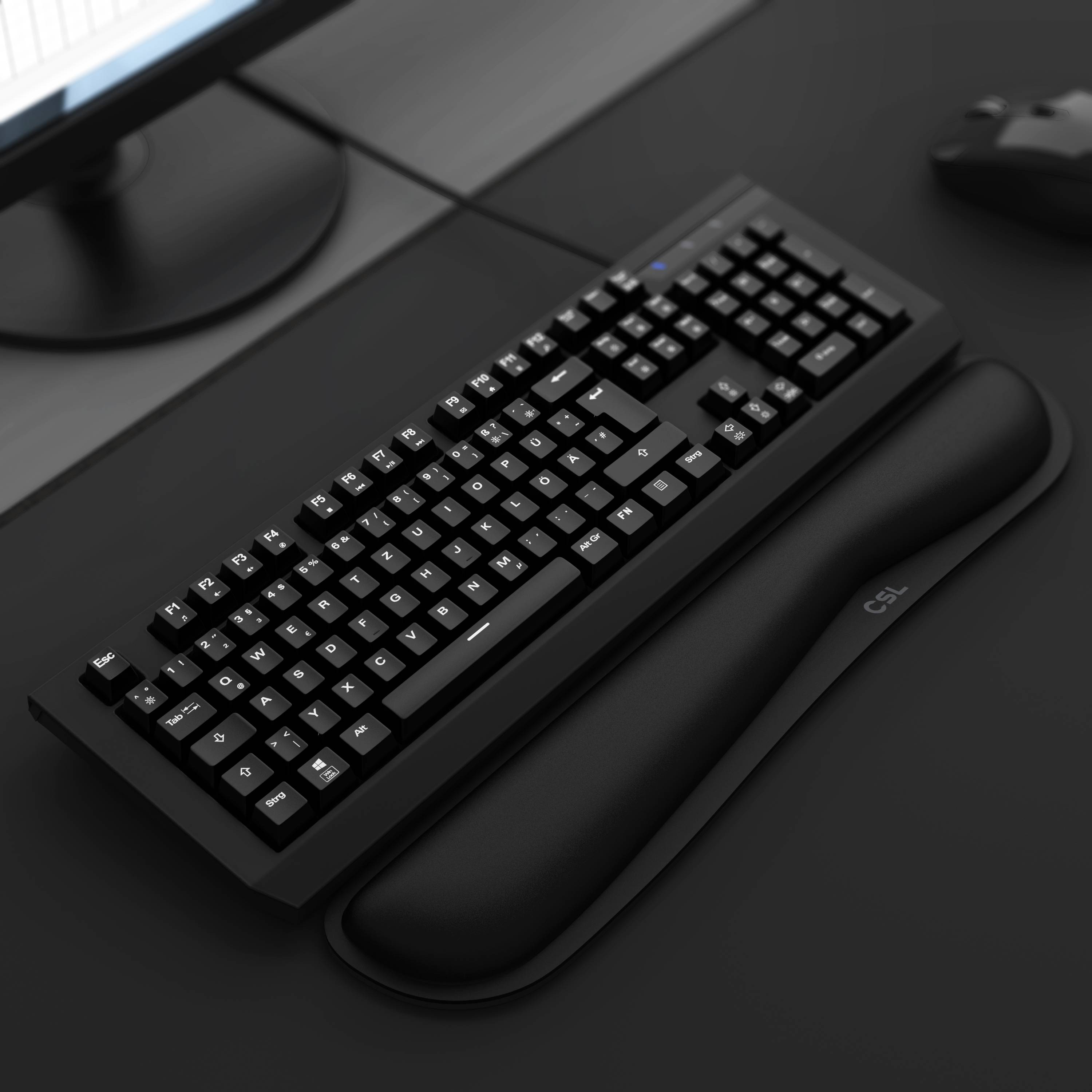 Schwarze mechanische Tastatur auf einem dunklen Schreibtisch mit Handgelenk­auflage und Teilen eines Monitors und einer Maus, die sichtbar sind.