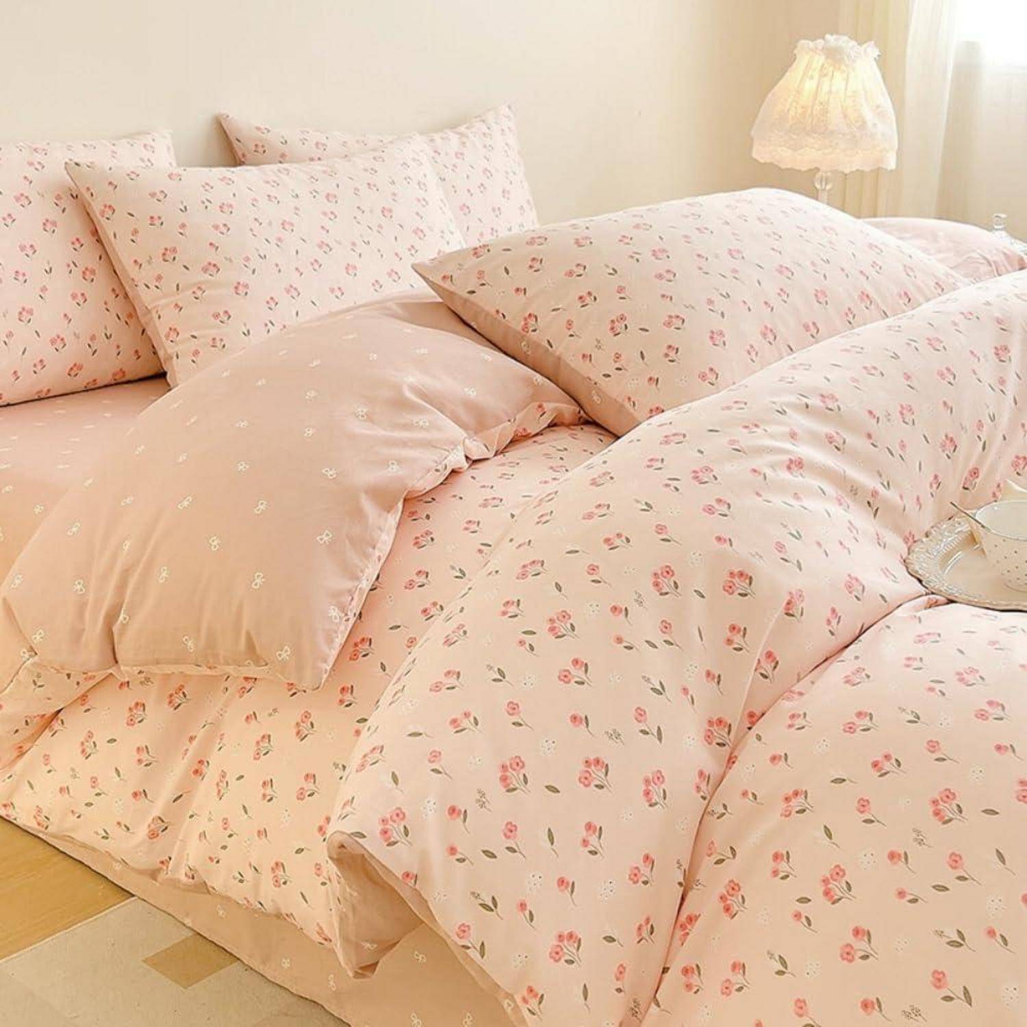 Ein Bett mit zarter rosa Blumenbettzeug, passenden Kissen und einer weichen cremefarbenen Lampe in einem hellen Schlafzimmer.