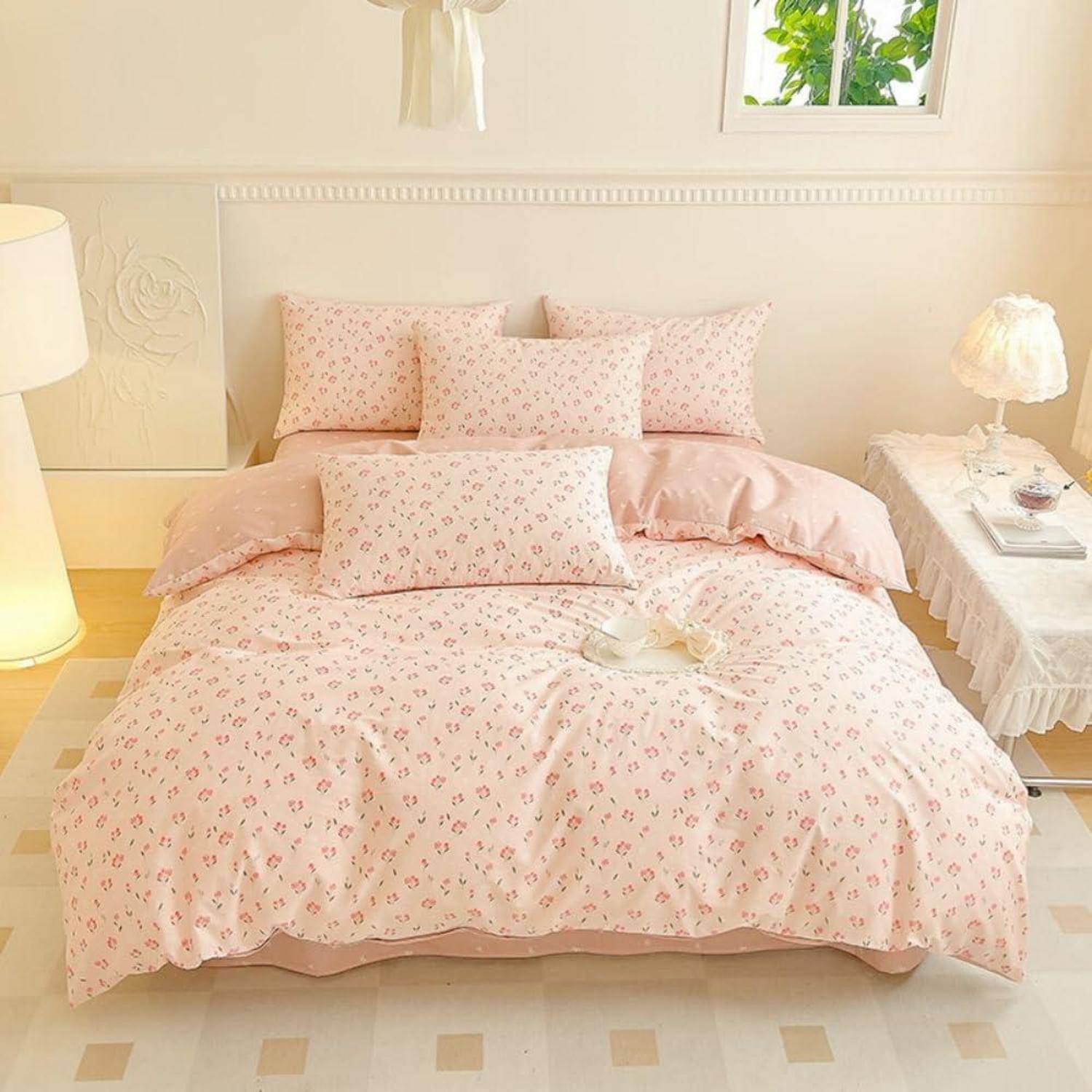 Zartes Rosa Blumenmuster-Bettwäsche auf einem ordentlich gemachten Bett in einem hellen, gemütlichen Schlafzimmer, mit Kissen, Nachttischlampen und einem kleinen Fenster.