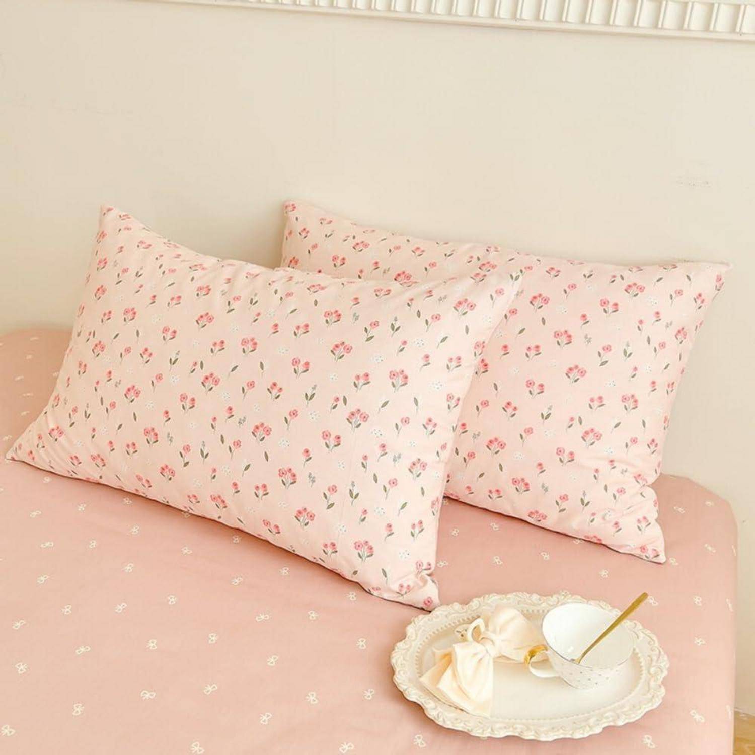 Zwei rosafarbene Blumenkissen auf einer pastellrosa Bettdecke, mit einem Spitzentablett, das eine weiße Tasse und ein gefaltetes Tuch im Vordergrund hält.