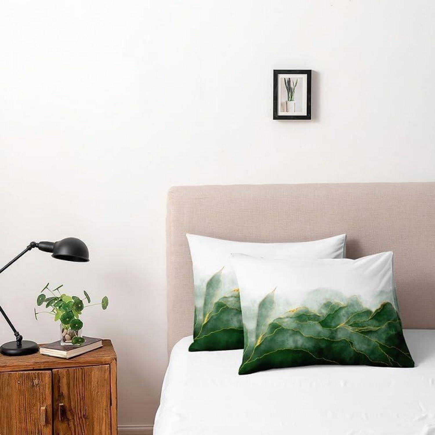 Ordentlich gemachtes Bett mit beigem Kopfteil, grünen Kissen mit Blattmuster, hölzernem Nachttisch mit Lampe und Pflanze sowie einem kleinen gerahmten Bild an einer weißen Wand.