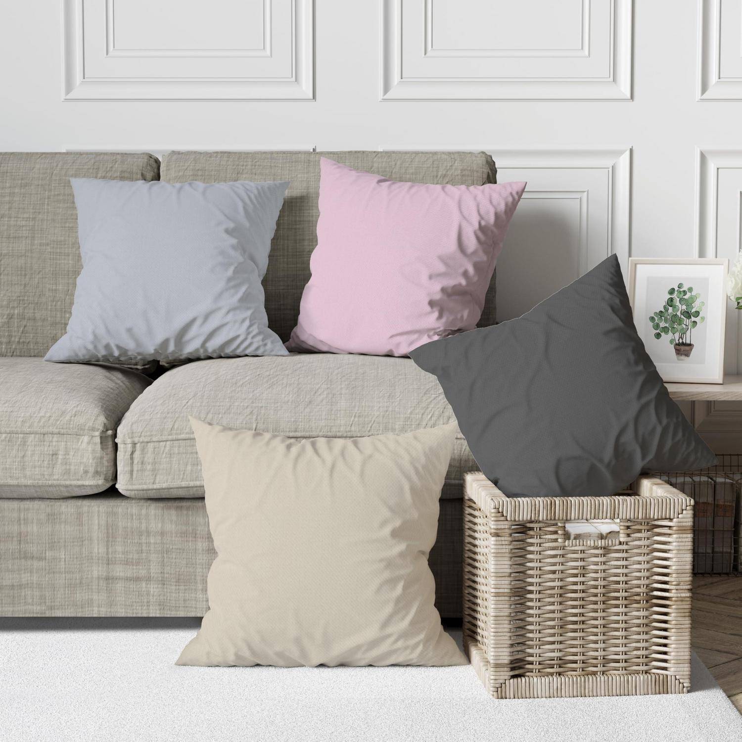 Vier quadratische Dekorationskissen in Hellgrau, Rosa, Dunkelgrau und Beige, die auf und neben einem neutralen Sofa in einem Wohnzimmer angeordnet sind.
