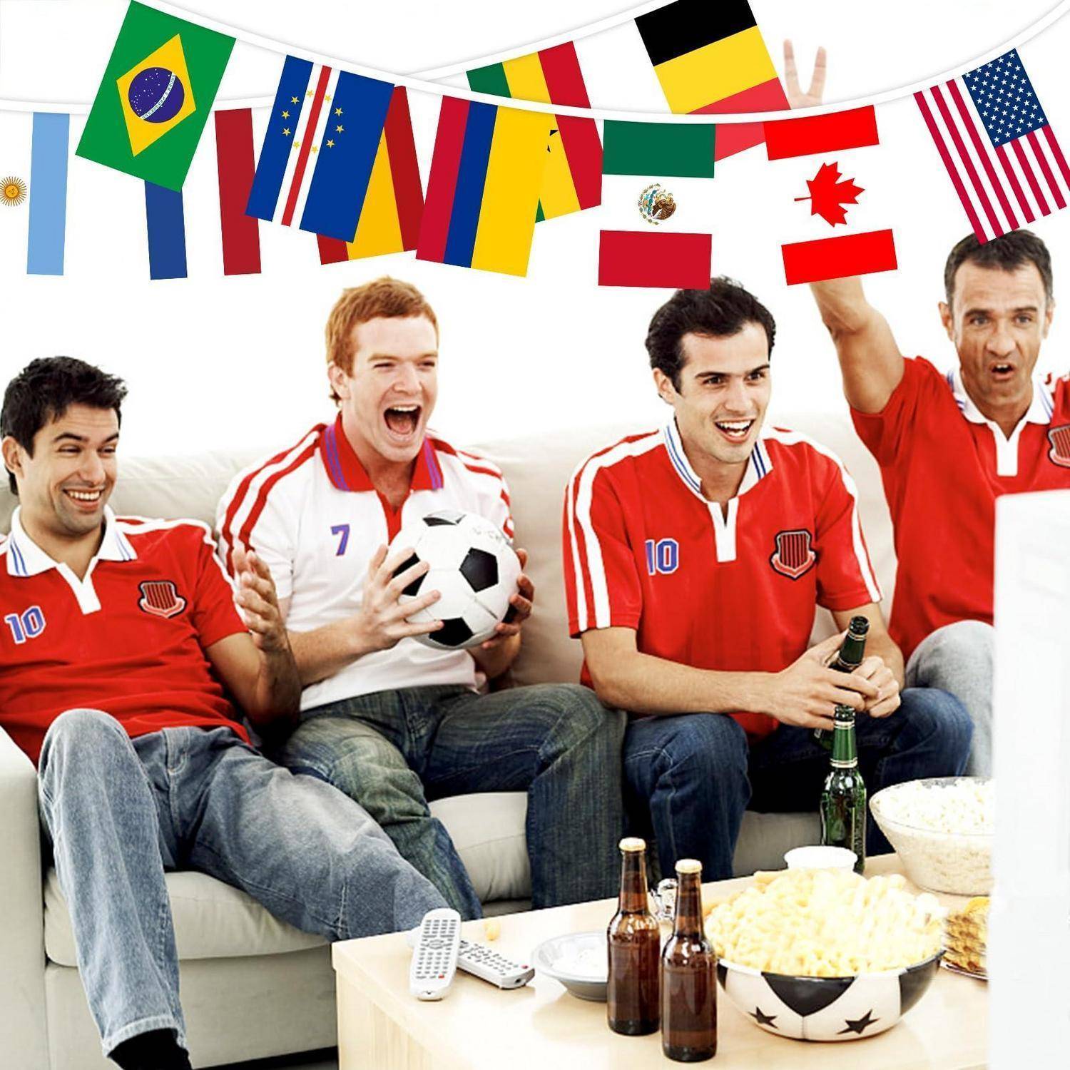 Vier Fußballfans in roten Trikots jubeln auf einem Sofa mit Getränken und Popcorn, während internationale Flaggen über ihnen hängen und eine Fußball-Weltmeisterschafts-Viewing-Party andeuten.