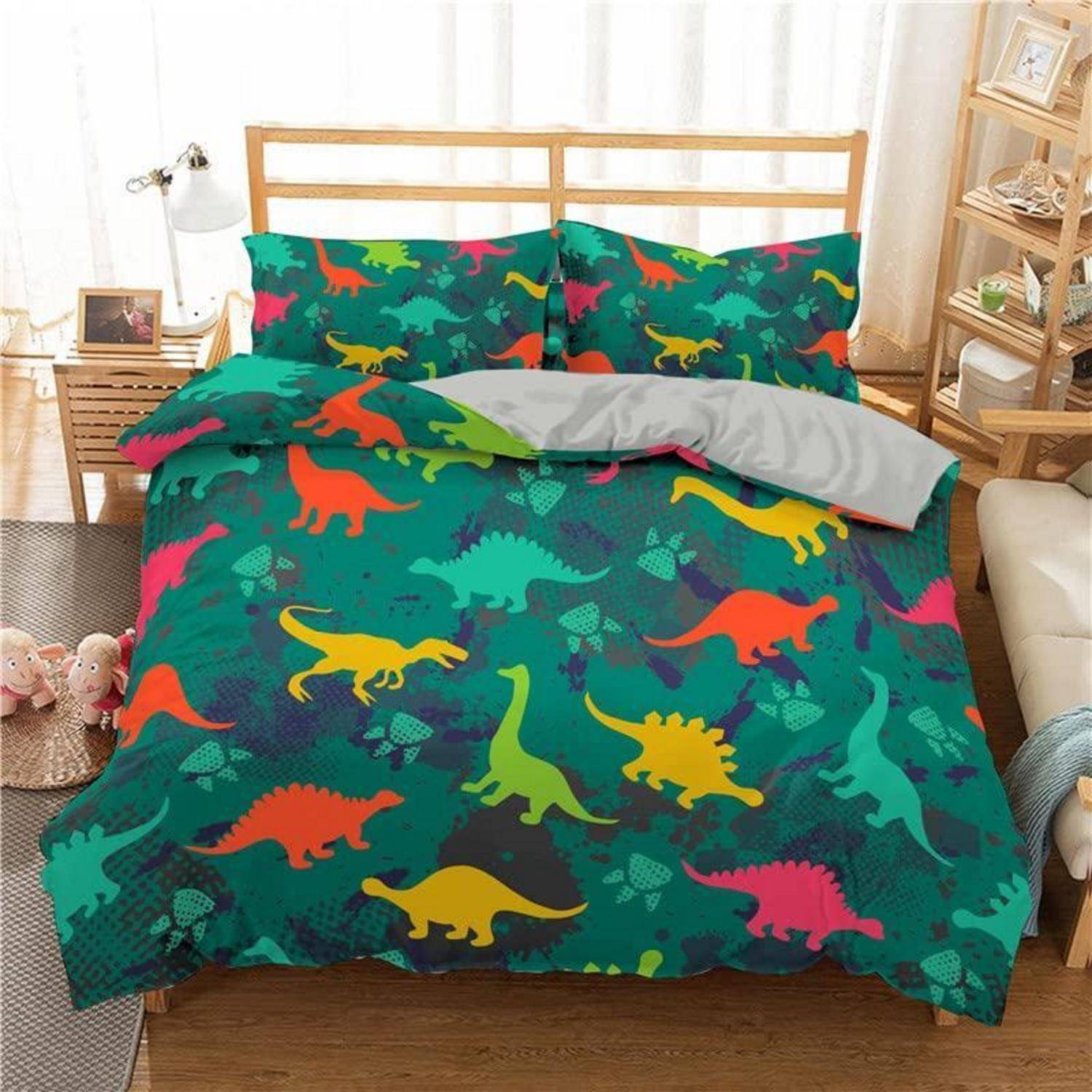 Bett mit buntem Dinosaurier-Muster-Bettwäsche und Kissen in einem hellen Schlafzimmer, mit einer gefalteten grauen Decke am Fußende des Bettes.