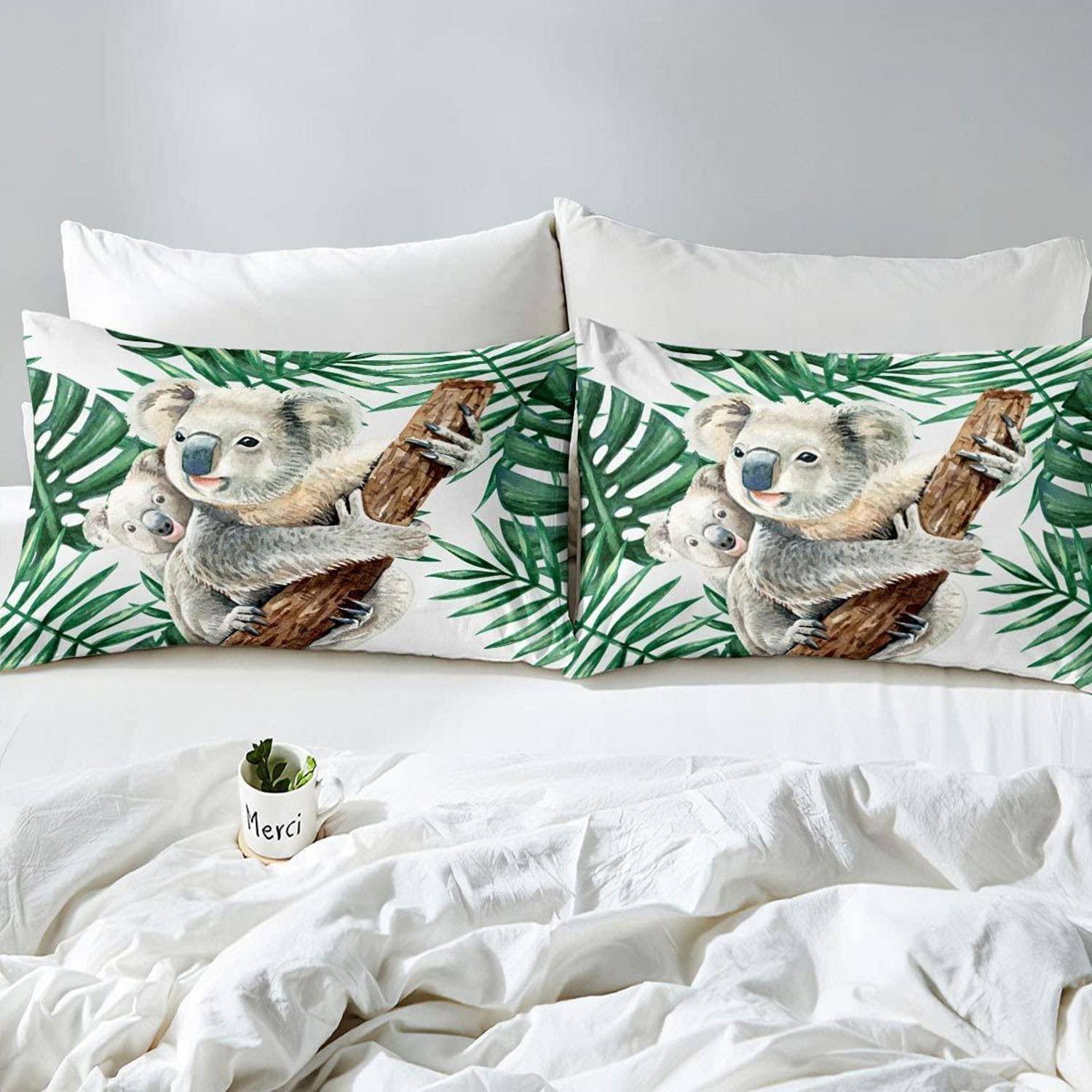 Zwei passende Kissen mit Koala-Muster auf einem weißen Bett mit weißer Bettwäsche und einer kleinen „Merci"-Tasse in der Nähe des Fußendes des Bettes.