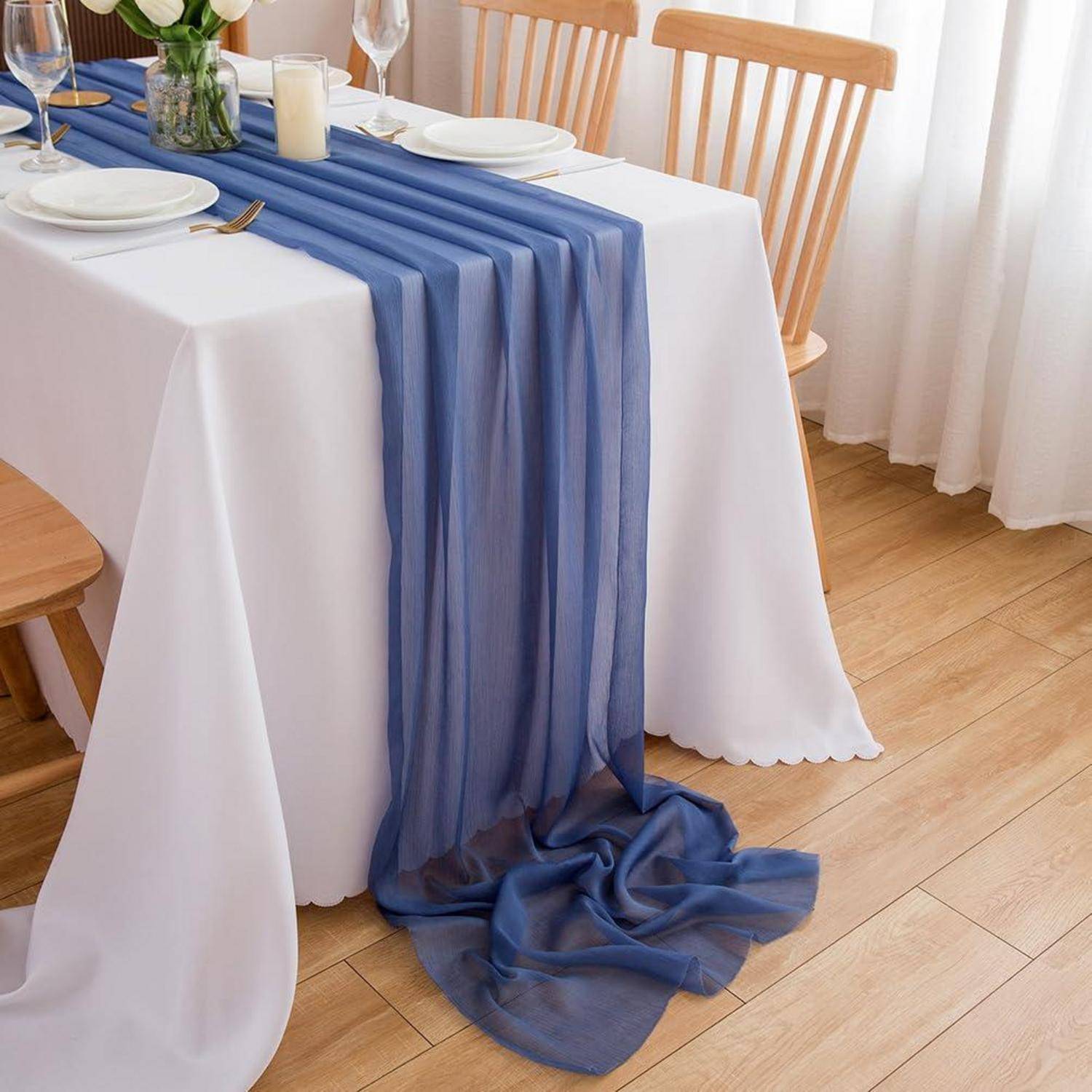 Esstisch mit weißer Tischdecke und blauem transparentem Tischläufer, eingedeckt mit Tellern und Gläsern in einem hellen Raum.