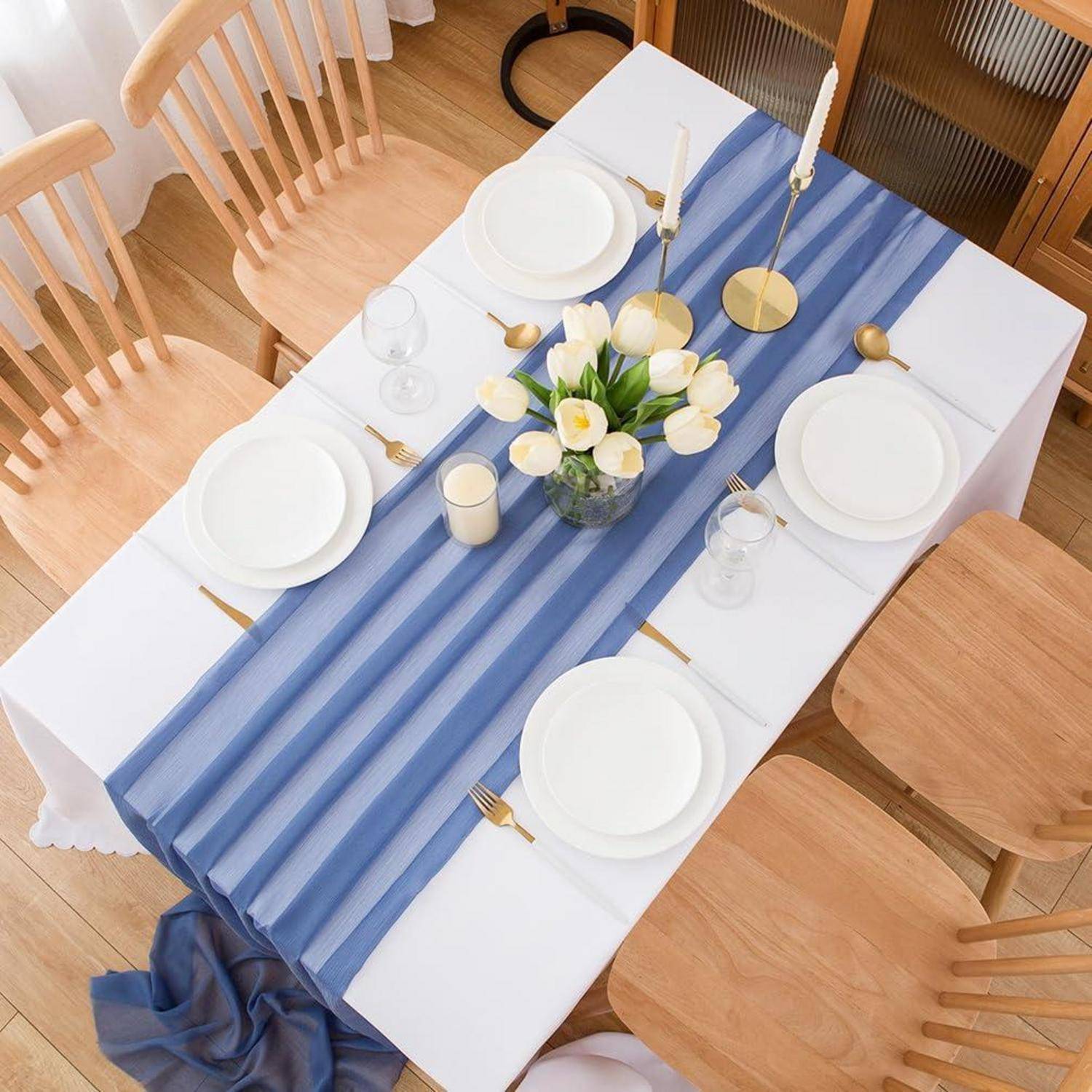 Draufsicht auf einen gedeckten Esstisch mit weißen Tellern, Kerzen, Glaswaren und einem blau gestreiften Läufer mit einem Blumenmittelstück.