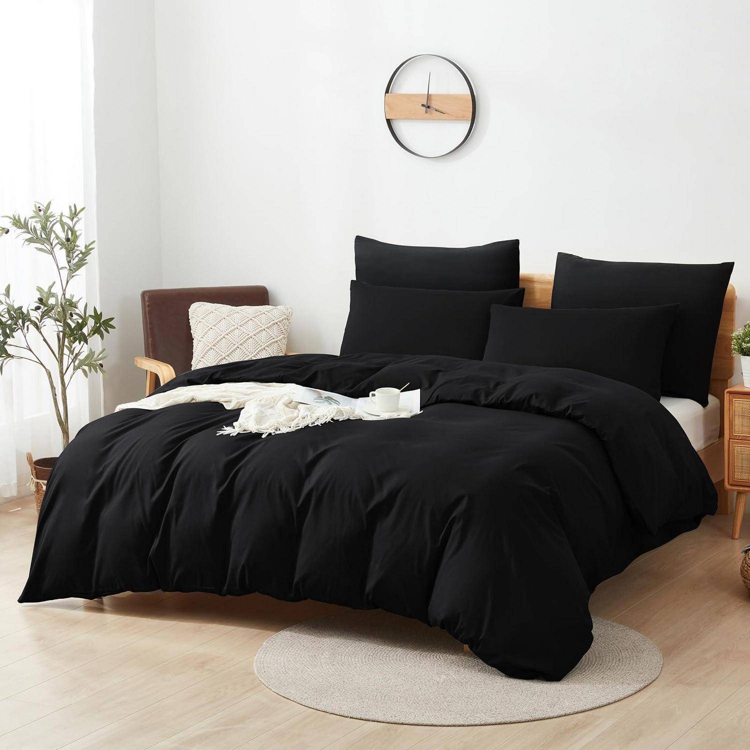Schwarze Bettwäsche auf einem gemachten Bett in einem hellen, minimalistischen Schlafzimmer mit Holzmöbeln, einer Wanduhr und einer Topfpflanze.