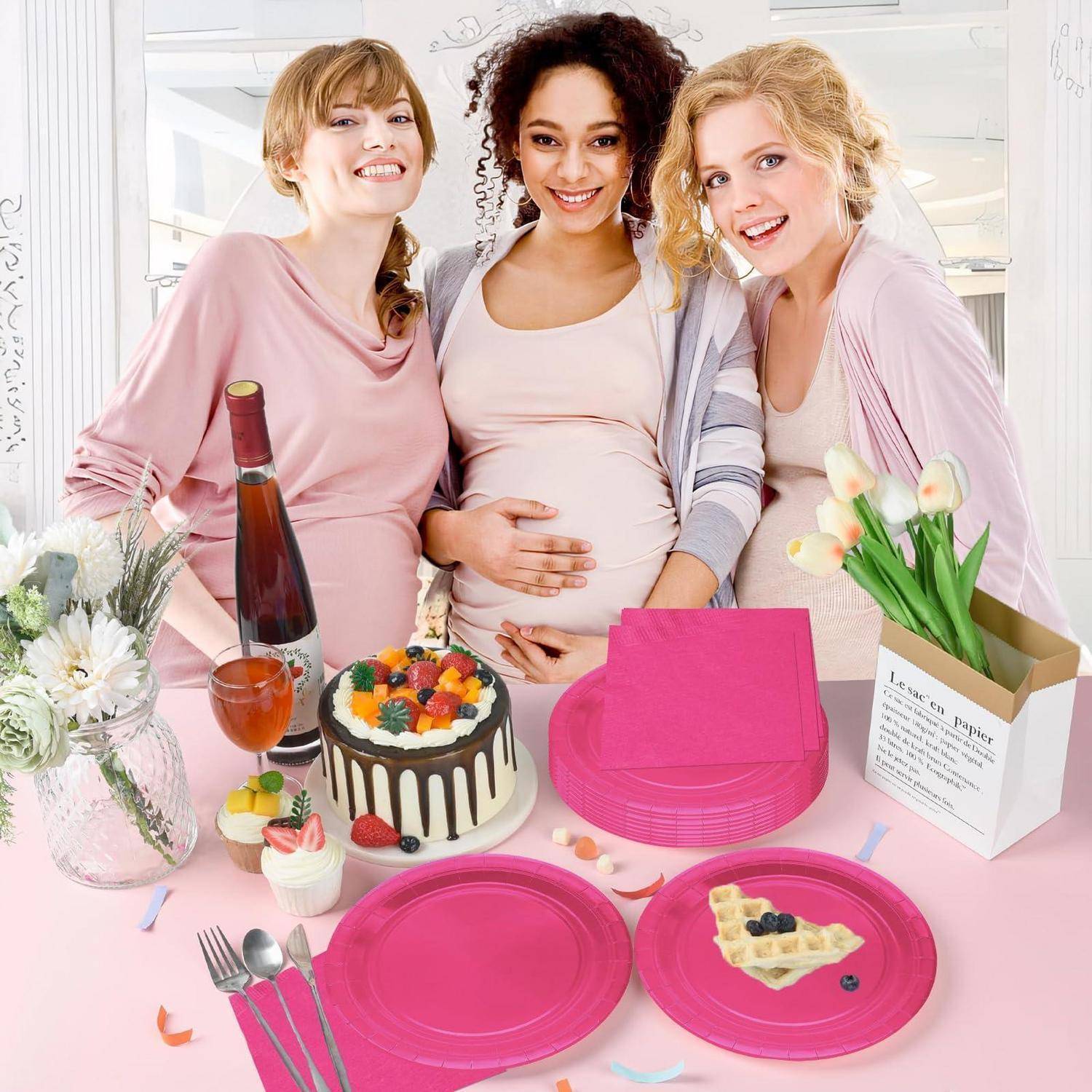 Drei schwangere Frauen lächeln an einem Babyduschtisch mit Kuchen, Geschenken, Blumen und rosafarbener Tischdecke.