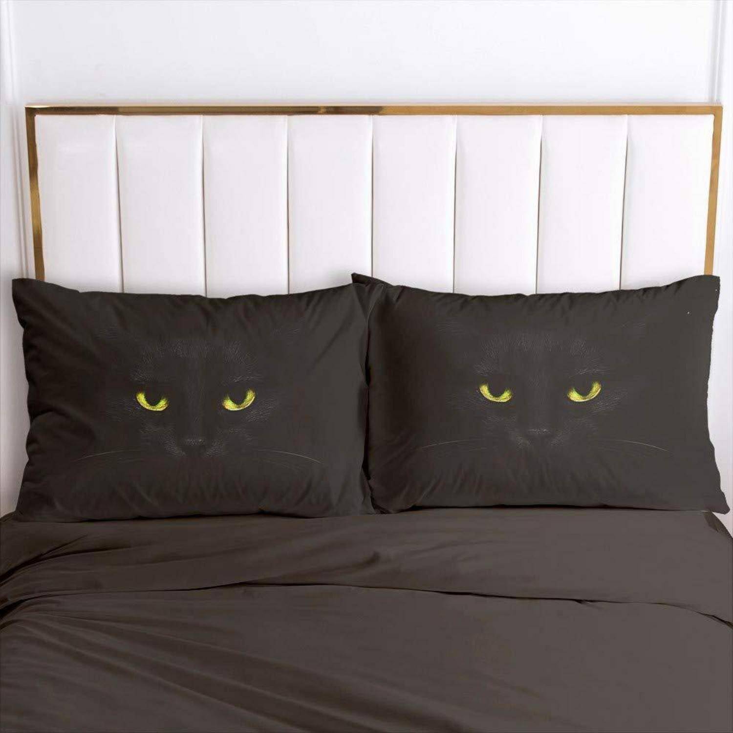 Zwei schwarze Katzenkopfkissen mit gelben Augen sitzen auf einem Bett mit dunkler Bettwäsche und einem weiß gepolsterten Kopfteil.