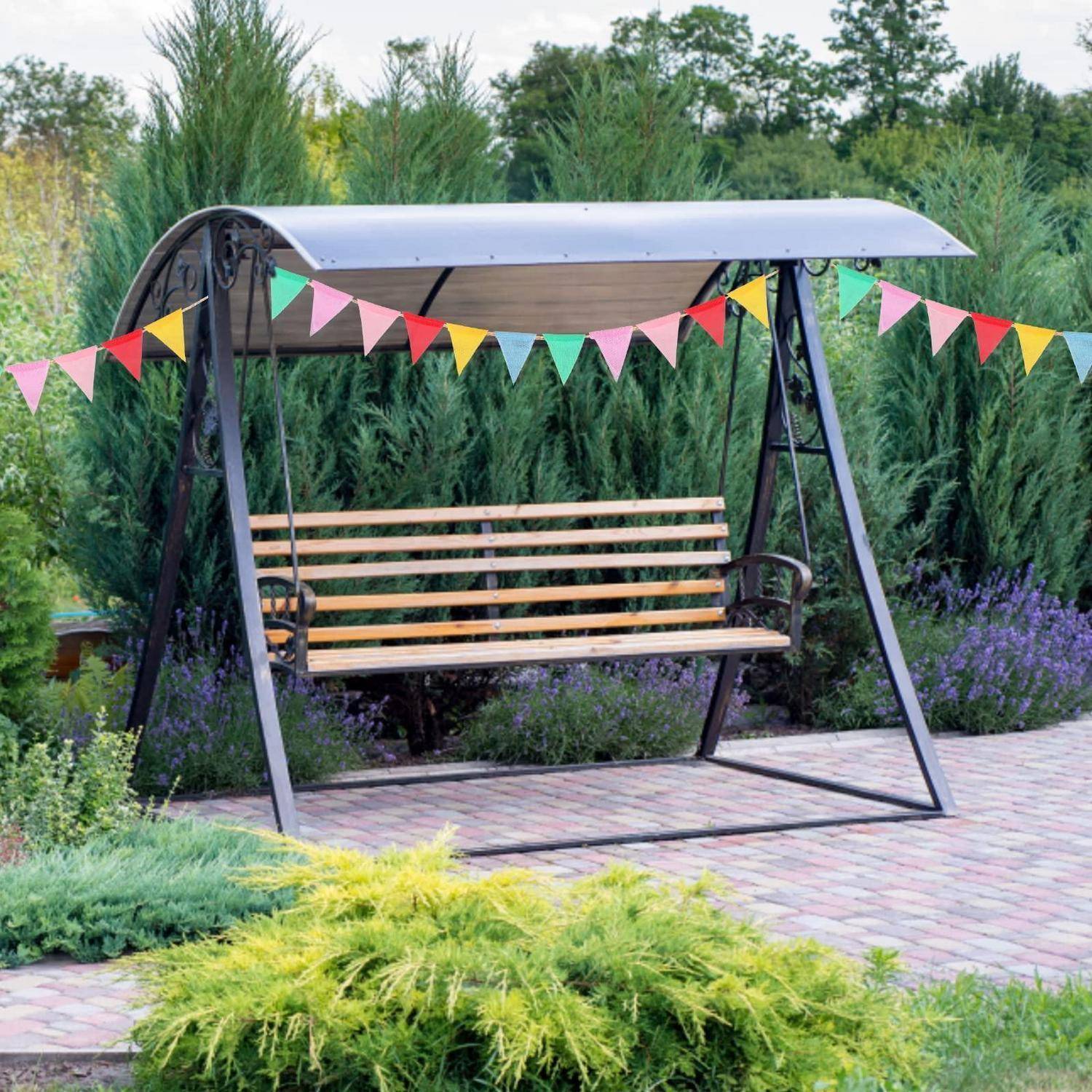 Gartenschaukel-Bank mit hölzernem Sitz und buntem Wimpel, auf einer gepflasterten Terrasse inmitten von üppiger Vegetation und Blumen.