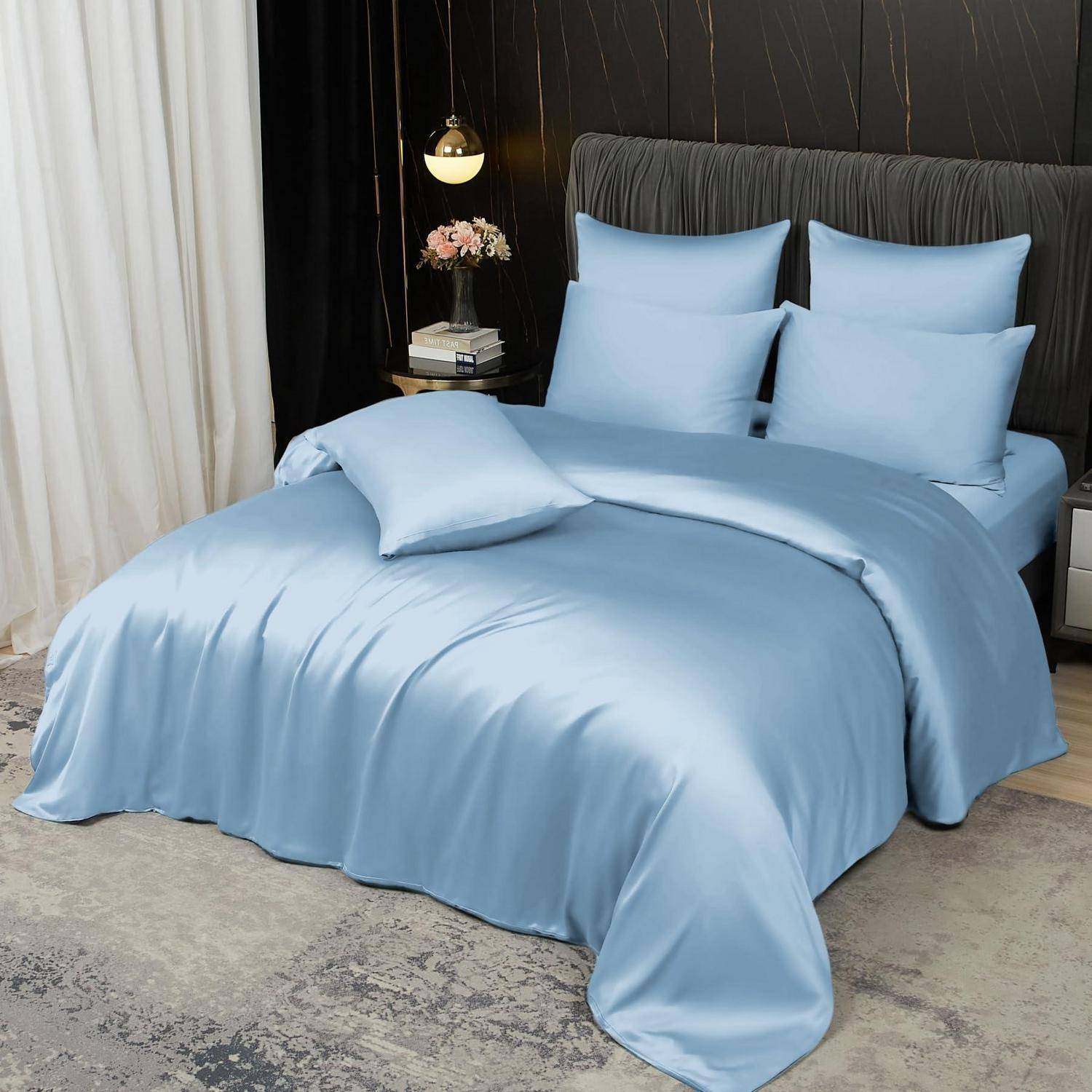 Hellblaubelur Satinbettwäsche auf einem ordentlich gemachten Bett mit passenden Kissen in einem dunkel eingerichteten modernen Schlafzimmer neben einer Lampe und einem Nachttisch.