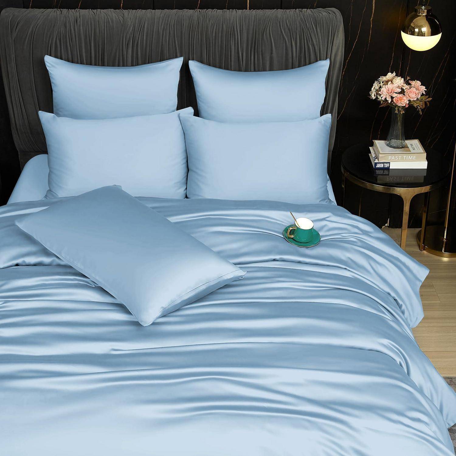 Bett mit hellblauem Satinbettzeug, mehreren Kissen und einem passenden dekorativen Wurfkissen in einem dunklen, modernen Schlafzimmer.
