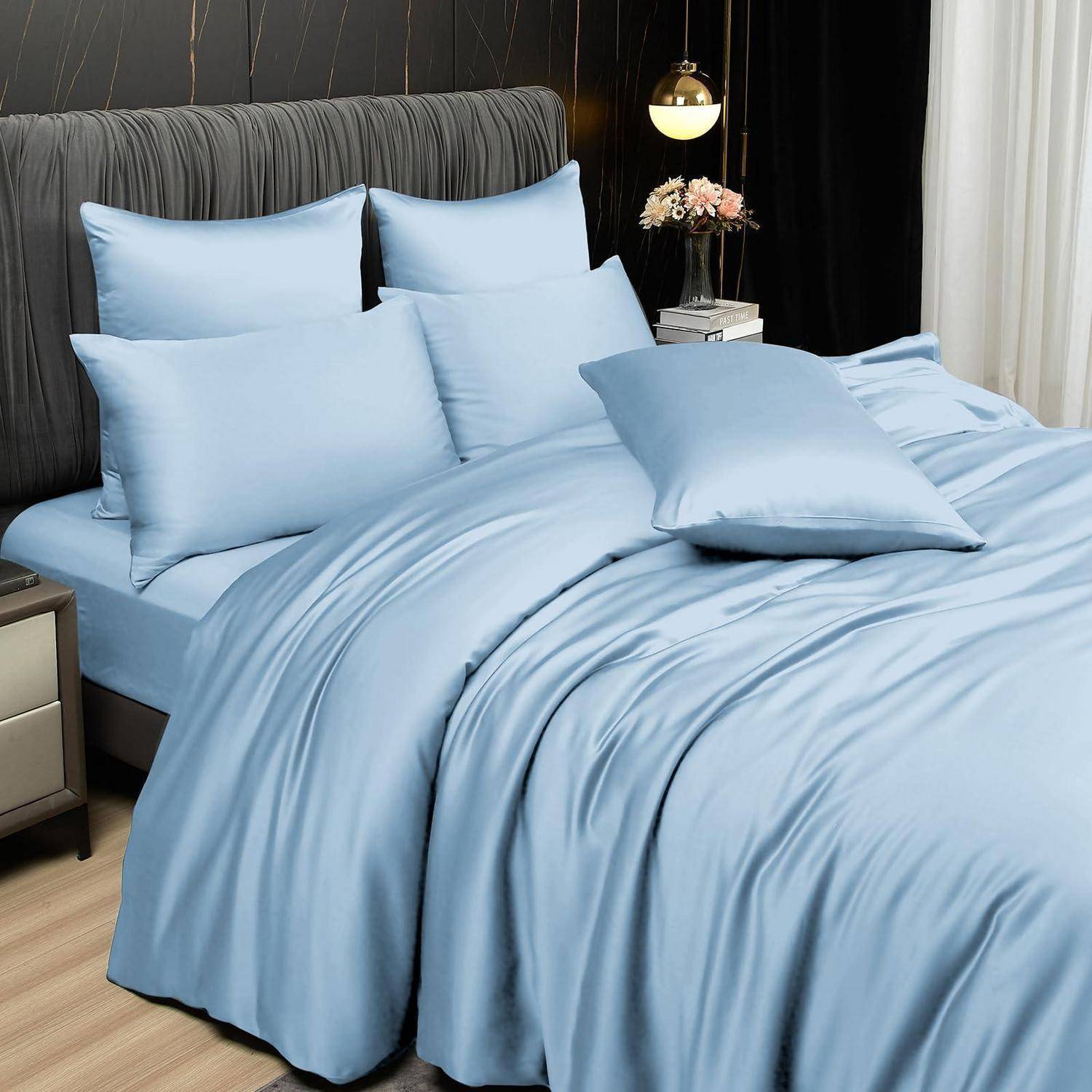 Ein sauber gemachtes Bett mit hellblauer Bettwäsche und mehreren Kissen in einem modernen Schlafzimmer, mit einer Nachttischlampe und Blumen daneben.