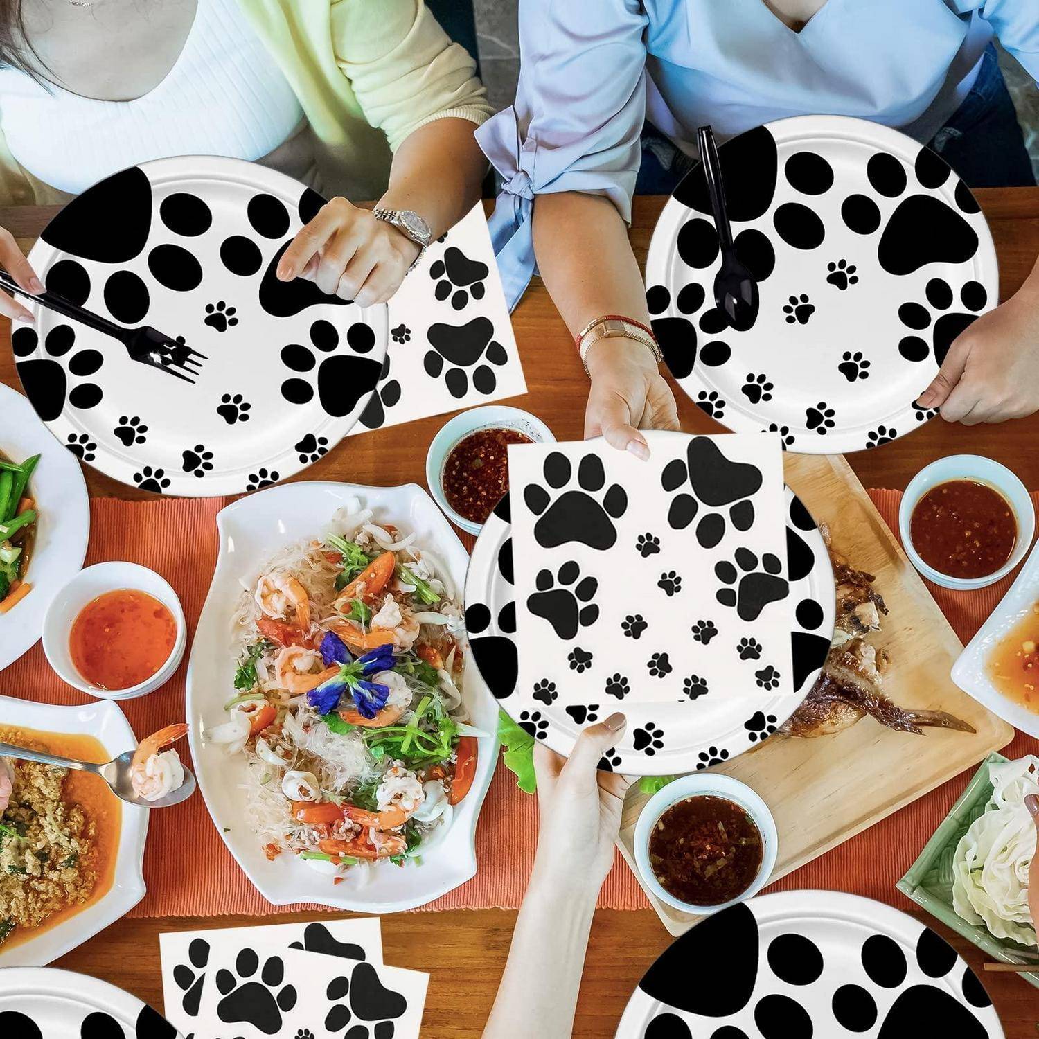 Menschen an einem Tisch halten weiße Teller mit schwarzen Pfotenabdrücken um gemeinsame Gerichte herum, was auf ein haustiergebendes Essen oder eine tiergebendes Event hindeutet.