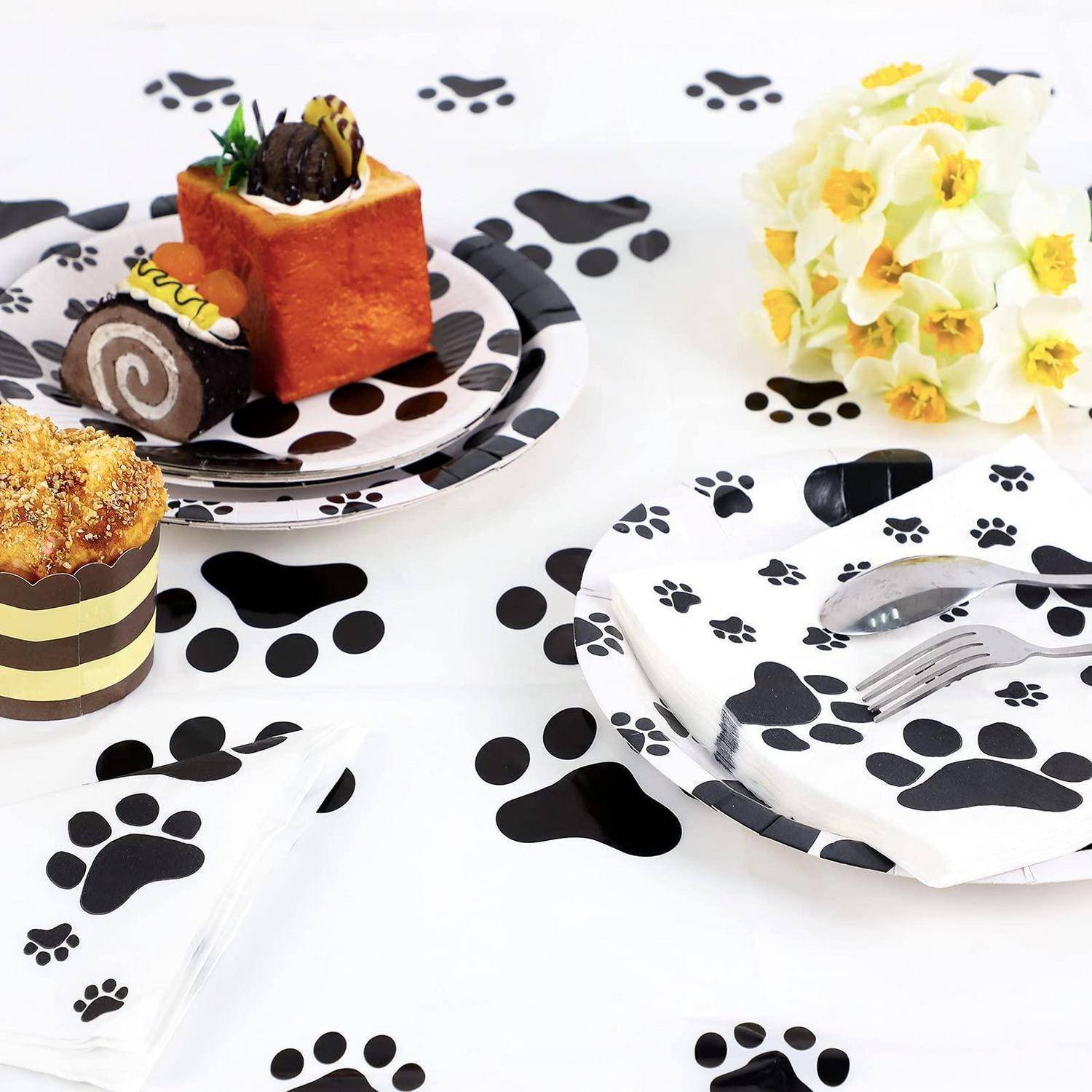Katzenthematische Tischdeckung mit Pfotendruck-Tellern, Servietten und Besteck, plus verschiedene Backwaren und einen Strauß aus weißgelben Blumen.