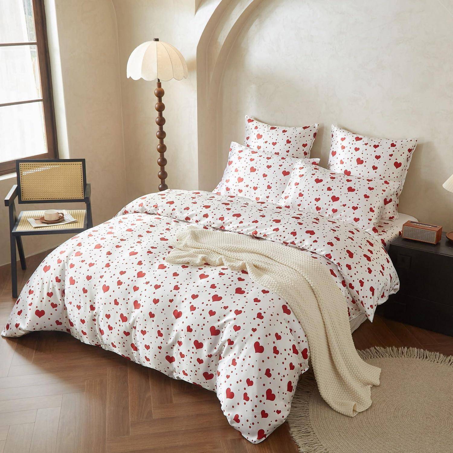 Ein ordentlich gemachtes Bett mit weißer Bettwäsche, die mit kleinen roten Herzmustern bedeckt ist, eine cremefarbene Wurfdecke und eine gemütliche Schlafzimmereinrichtung mit einer Lampe und einem Stuhl.
