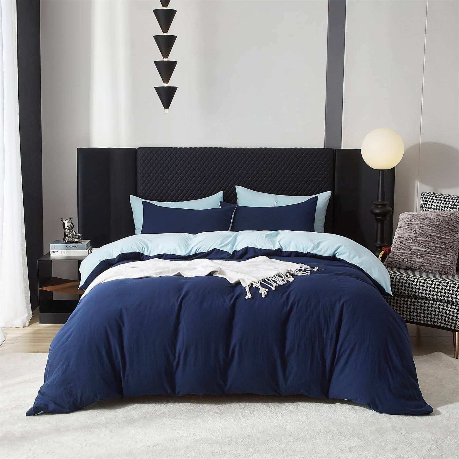 Ein ordentlich gemachtes Bett mit dunkelblauer und hellblauer Bettwäsche in einem modernen Schlafzimmer mit schwarzem Kopfteil, Nachttischlampen und grauem Akzentuhl.