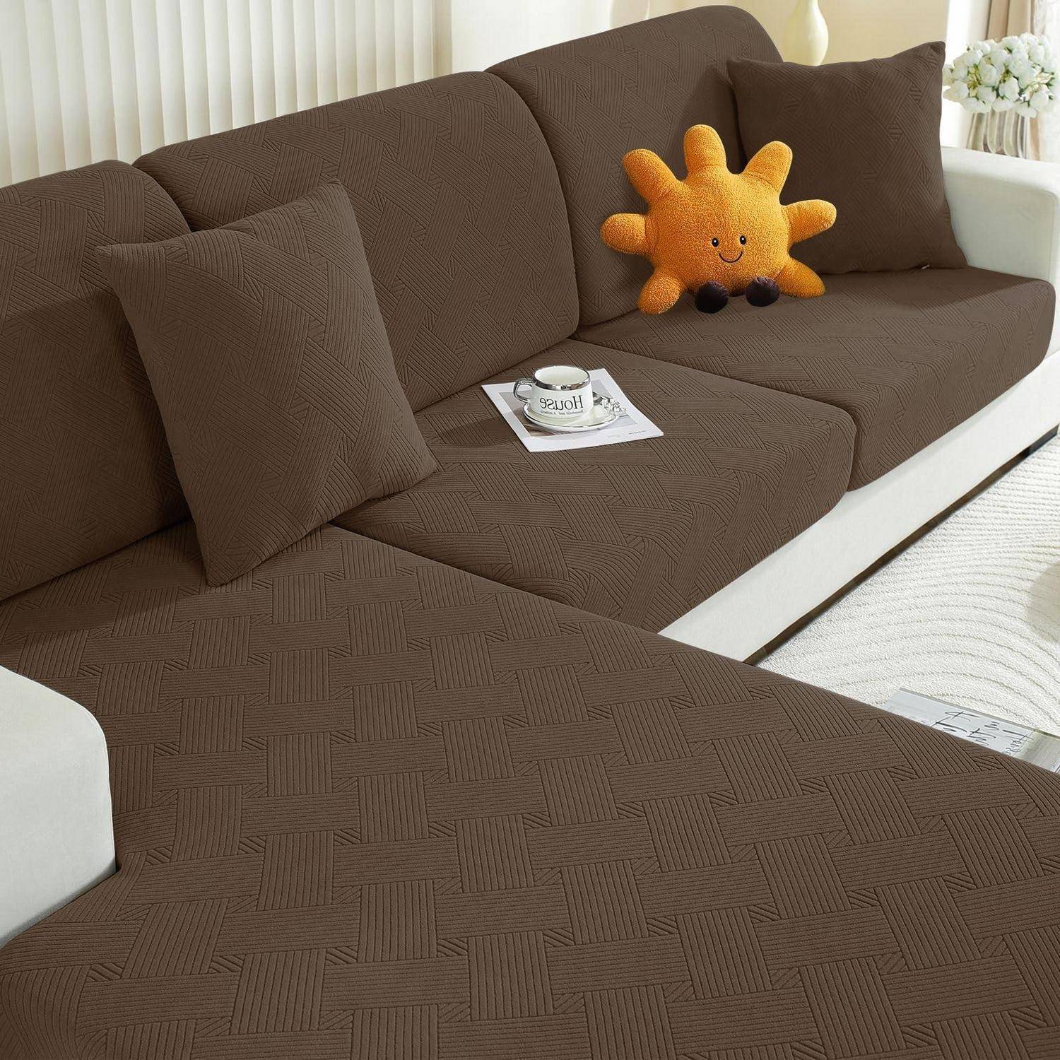 Braunes L-förmiges Sofa mit Schonbezug, zwei einfache Kissen, eine lächelnde orange Plüschsonne und eine Tasse auf einem Magazin in einem hellen Wohnzimmer.