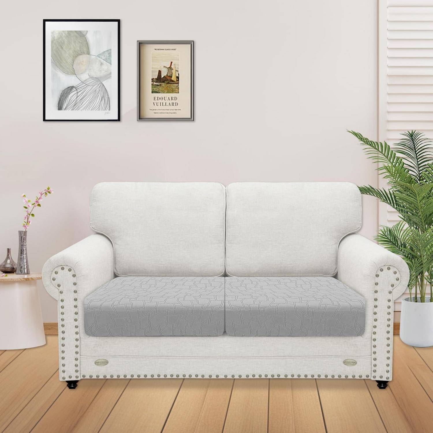 Hellgraues Zweisitzer-Sofa mit nietenbesetzten Armlehnen in einem hellen Wohnzimmer, an einer Wand mit gerahmter Kunst, einem Beistelltisch und einer Topfpflanze.