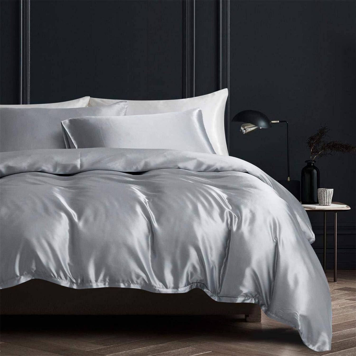 Ein ordentlich gemachtes Bett mit silbergrauer Satinbettwäsche und Kissen in einem dunklen, modernen Schlafzimmer neben einer kleinen Lampe und einem Nachttisch.