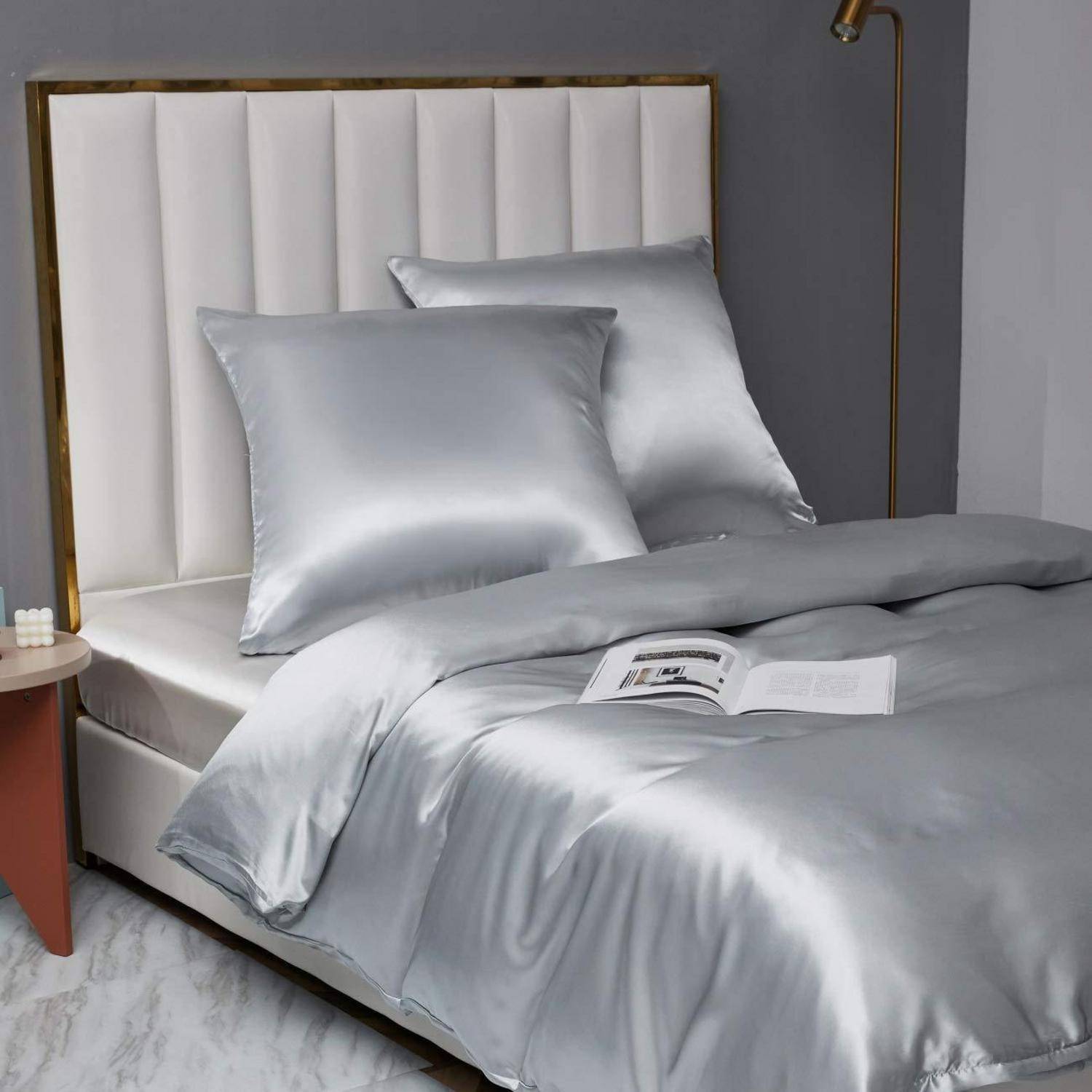 Ein ordentlich gemachtes Bett mit silbernem Satinbettzeug und zwei passenden Kissen, mit einem offenen Magazin darauf in einem modernen Schlafzimmer.