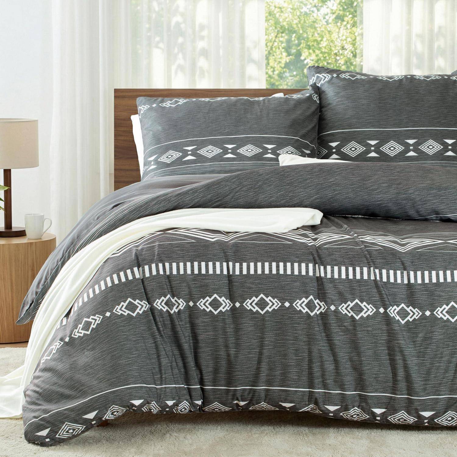 Ein ordentlich gemachtes Bett mit dunkelgrauem geometrischem Bettwäsche-Muster und weißen Laken in einem hellen Schlafzimmer mit einem hölzernen Nachttisch und einer Lampe.