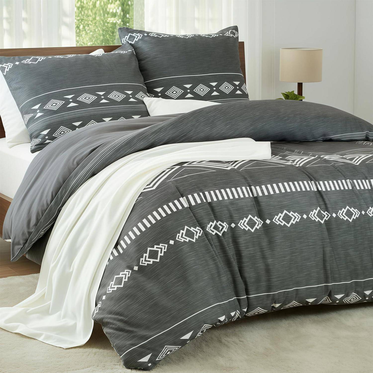 Ein ordentlich gemachtes Bett mit dunkelgrauer und weißer geometrischer Bettwäsche, weiße Laken über der Bettdecke drapiert, und passende Kissen in einem hellen Schlafzimmer.