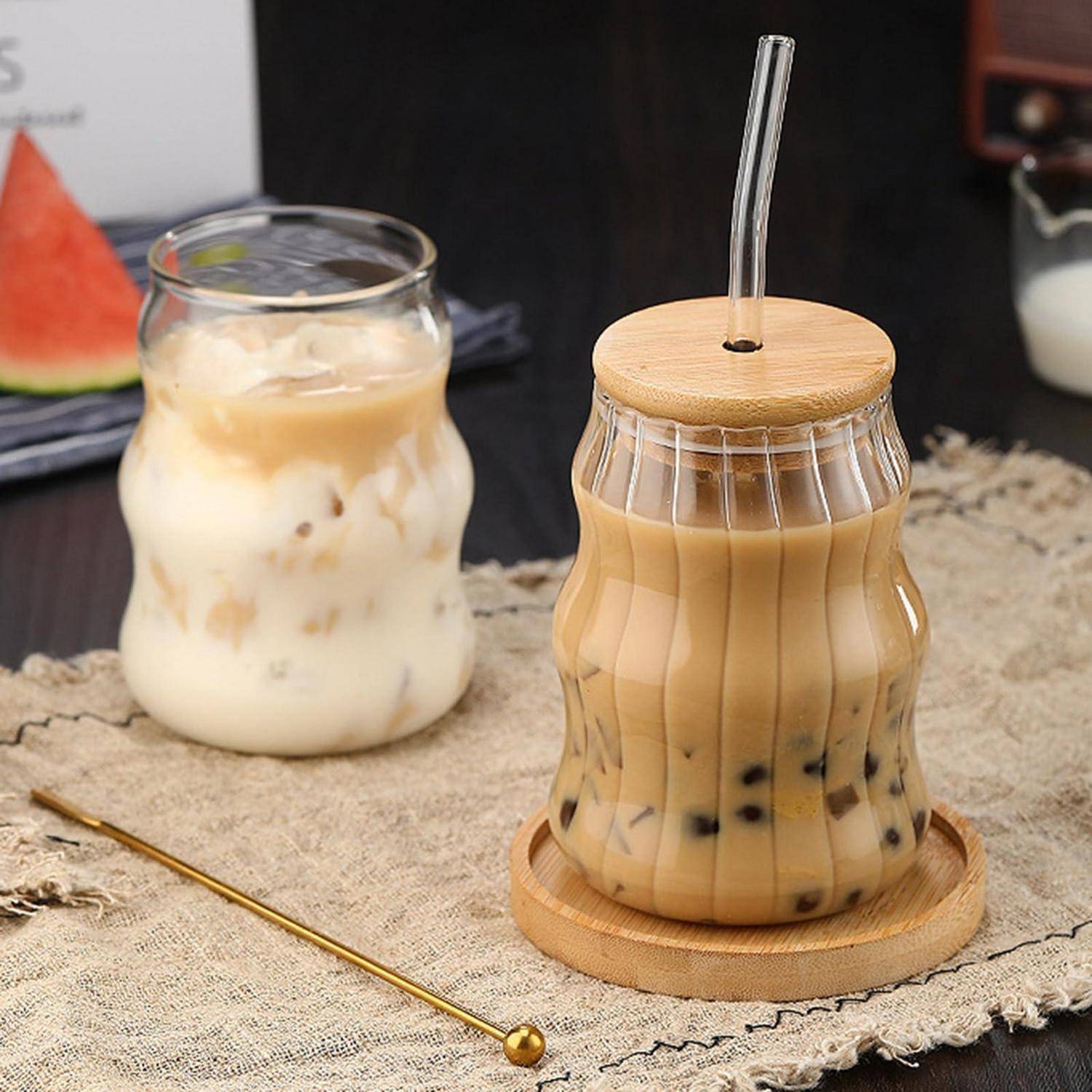 Zwei eisgekühlte Getränke in Glasgefäßen auf einem Tuch: ein cremiges Milchgetränk mit Eis und ein beiger Bubble Tea mit Bambusdeckel, Strohhalm und Untersetzer.
