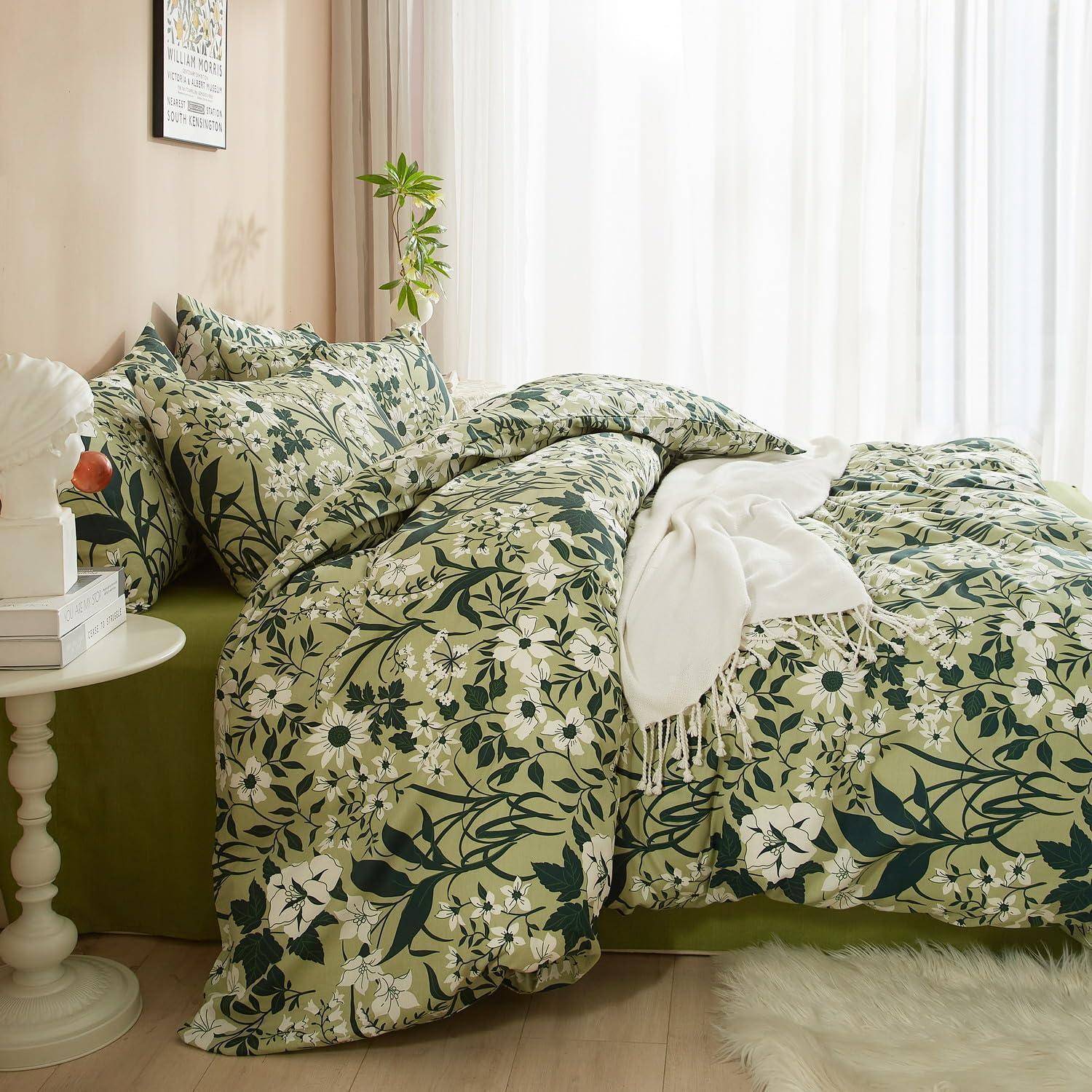 Blumig grüne Bettwäsche auf einem Bett in einem hellen Schlafzimmer, mit einer weißen Wurfdecke, Nachttisch, Pflanze und Vorhängen neben einem sonnenbeschienenen Fenster.