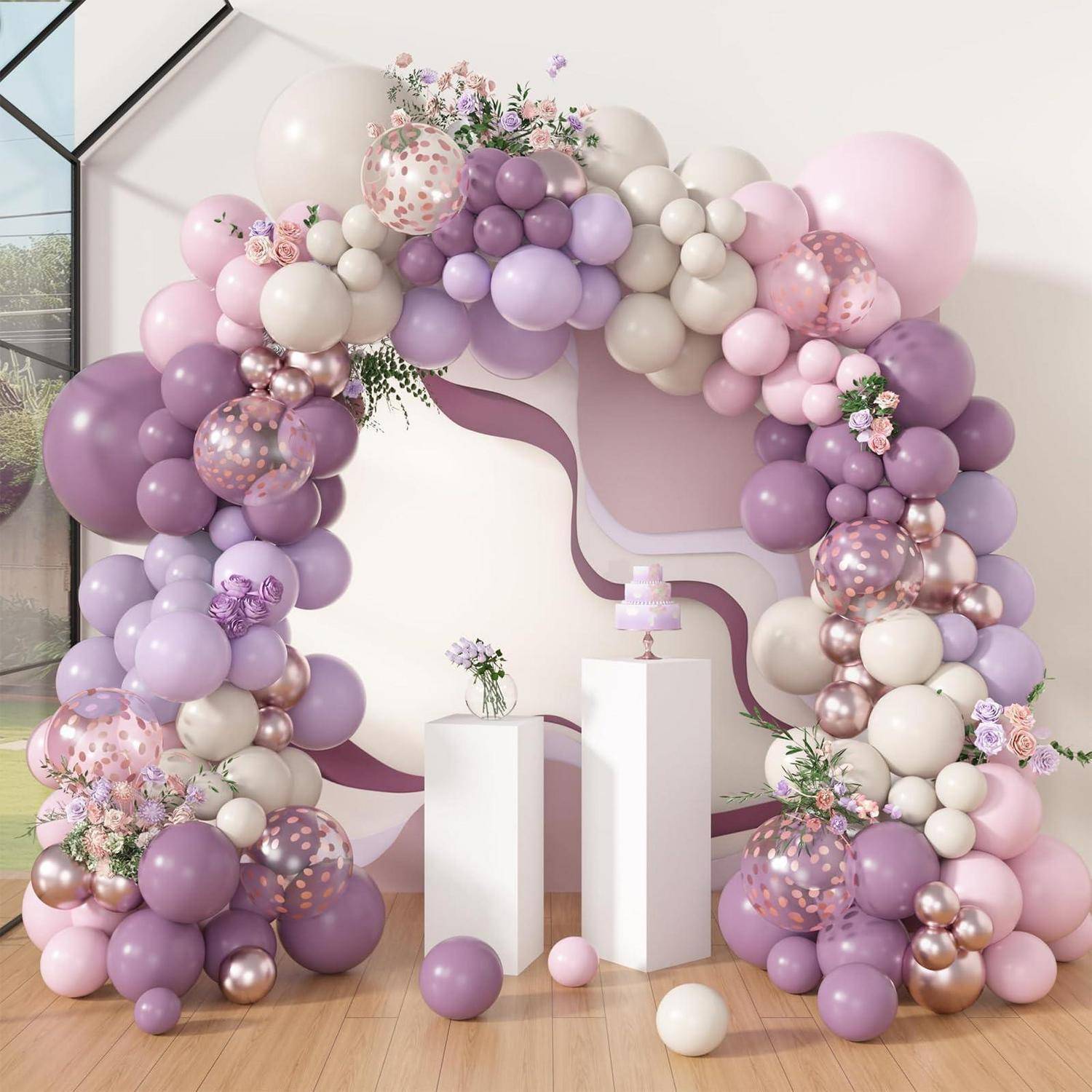Lavendel-, Rosa- und Creme-Ballongirlande mit Blumenakzenten rund um zwei weiße Sockel und kleine Kuchen für eine Partydekoration.